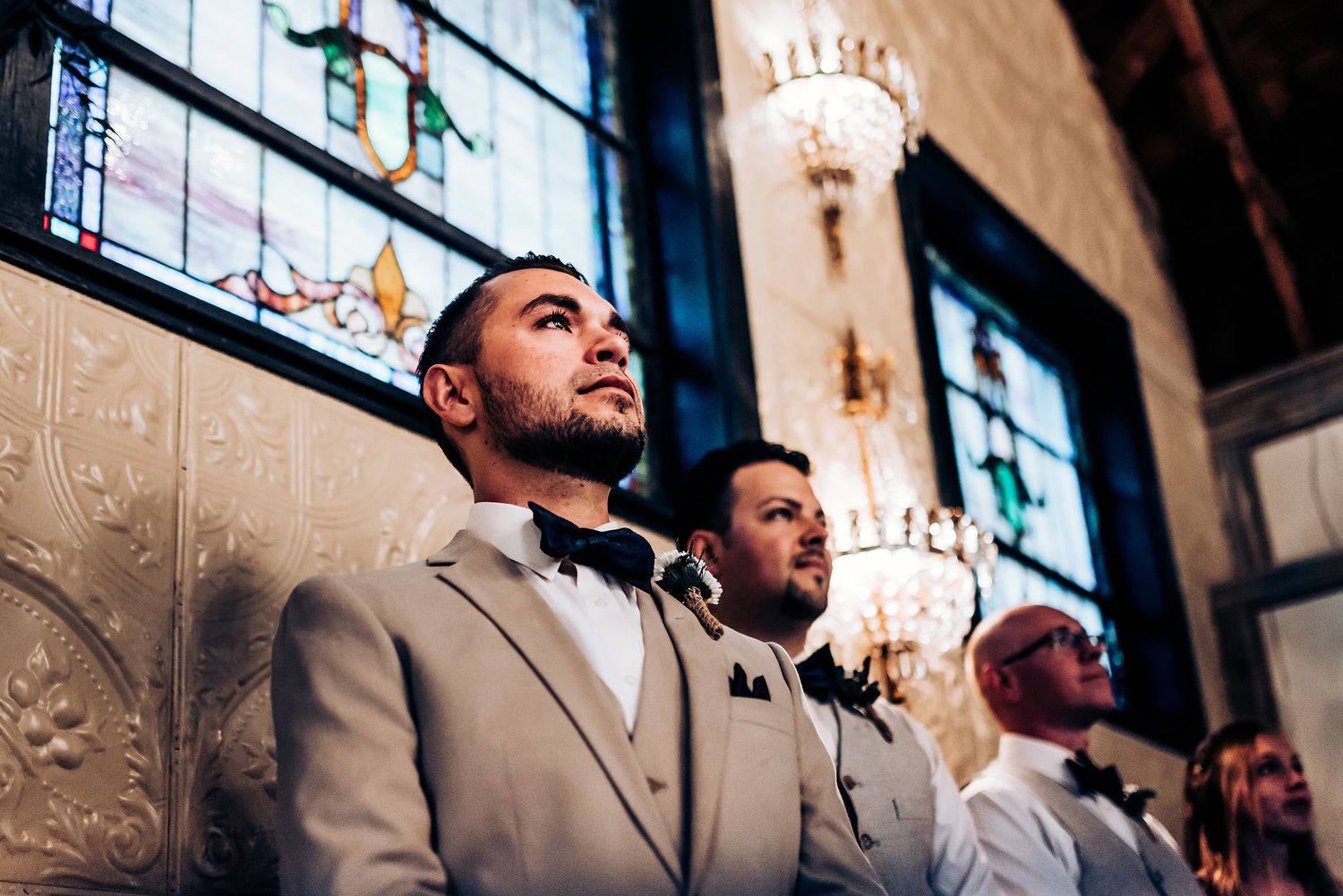 Groomsmen in formal attire stand beneath stained glass windows in an elegant venue.