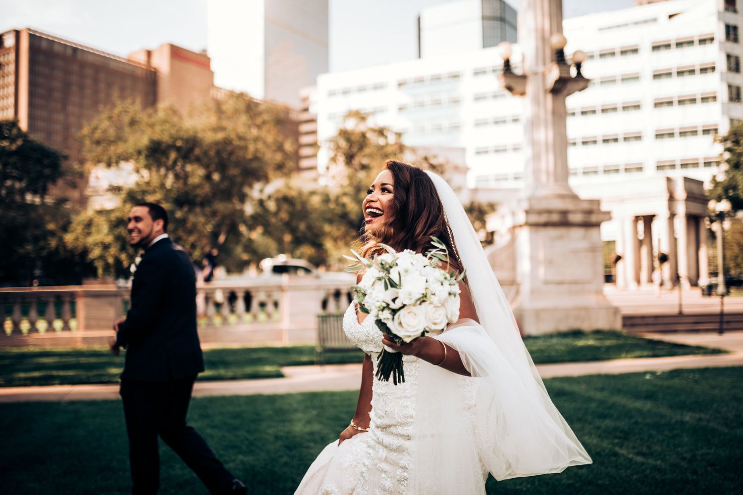 A bride smiles with bouquet in foreground while groom stands at distance in urban setting.