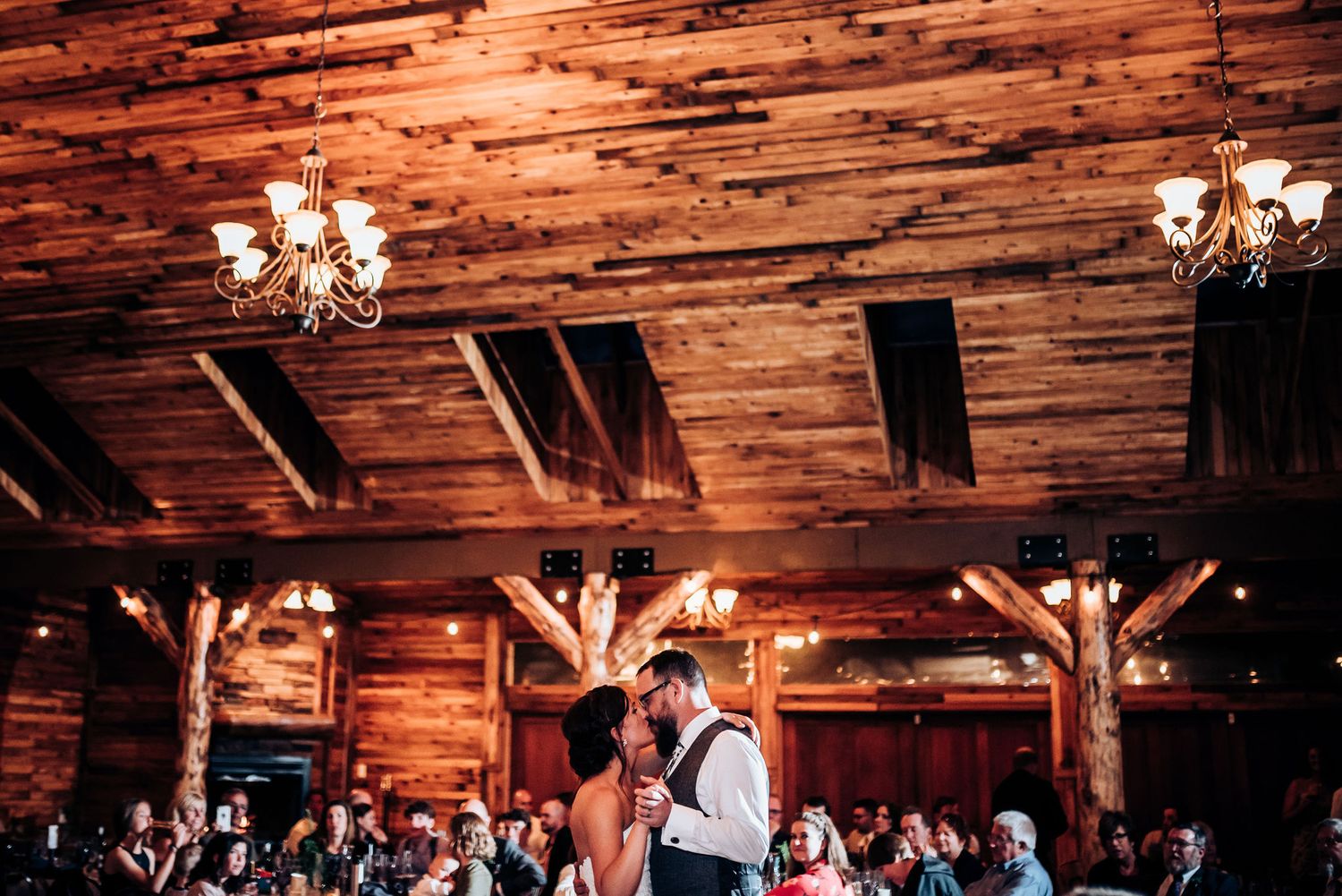 Wedding dance in a rustic wood-beamed venue with chandeliers and watching guests.