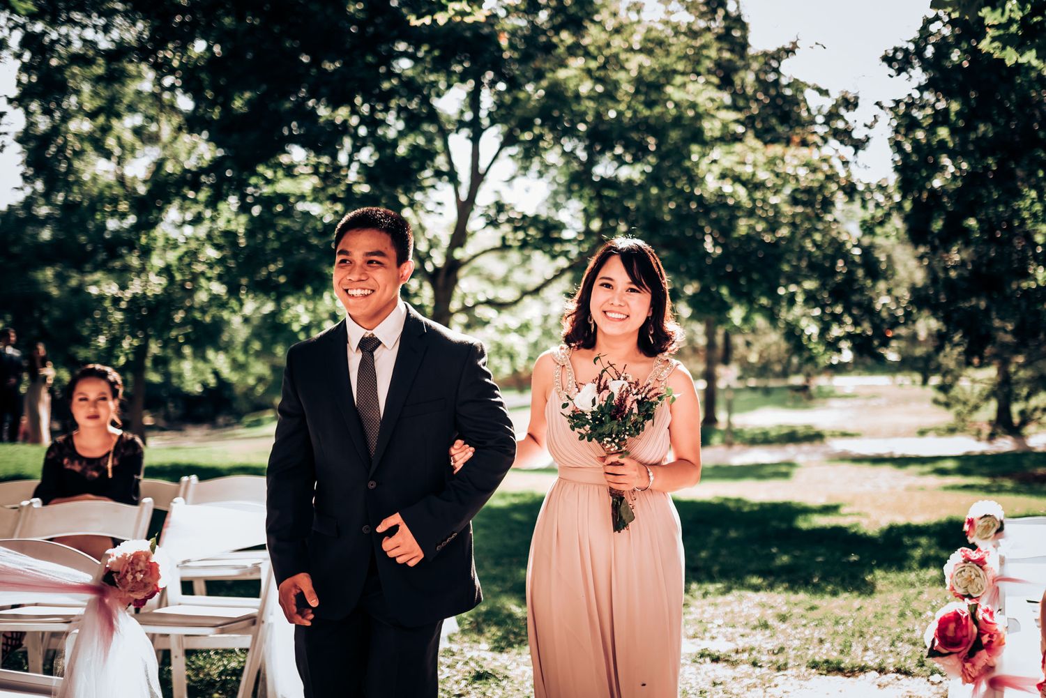 Wedding party members in formal attire walk down an outdoor ceremony aisle on a sunny day.
