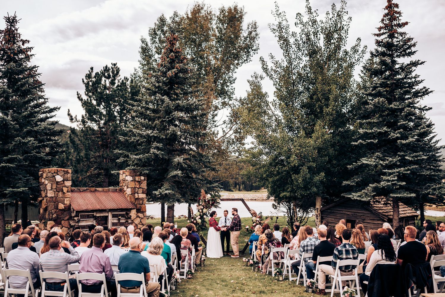 Guests seated in white chairs attend an outdoor wedding ceremony surrounded by tall evergreen trees.
