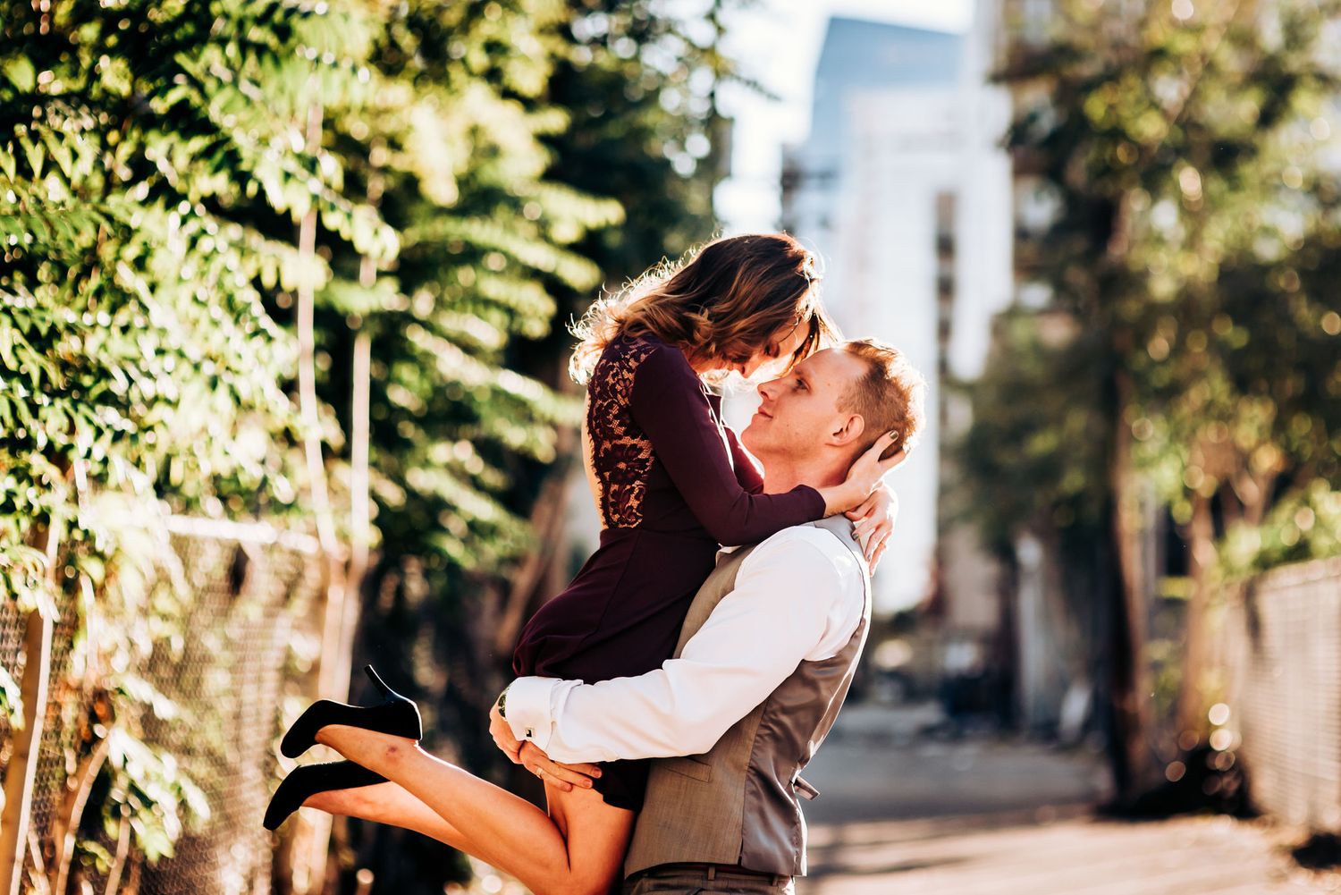 A romantic couple embraces passionately in an outdoor urban setting with greenery.