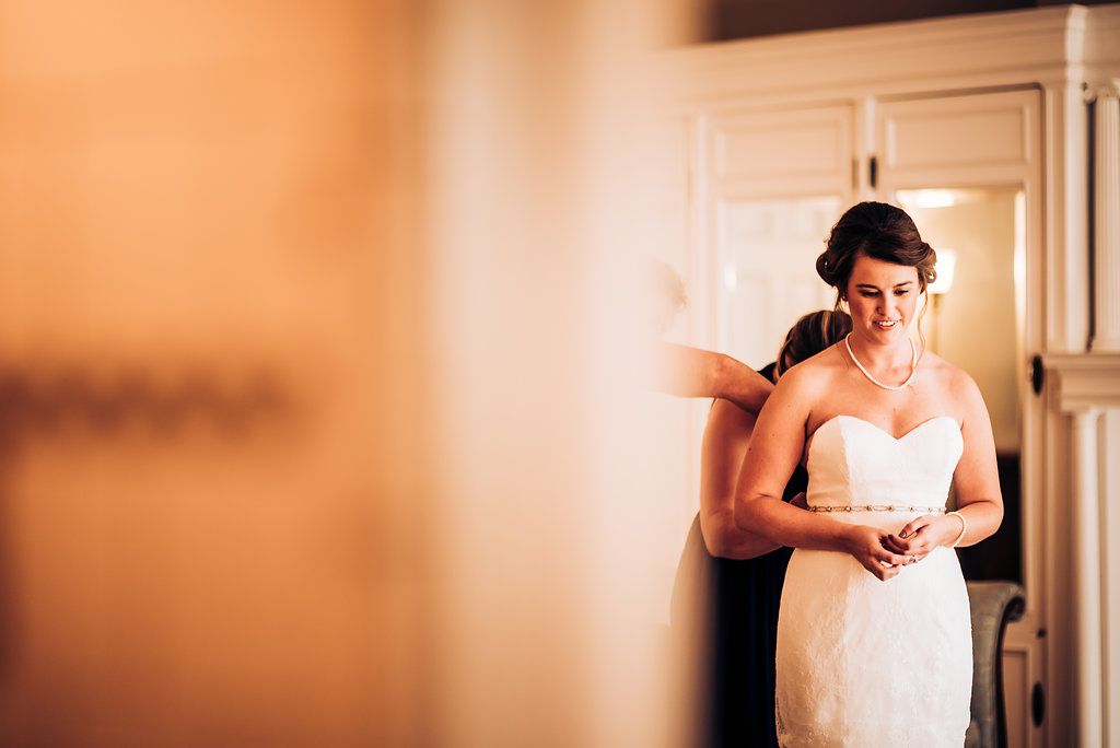 A bride in a white strapless dress stands near a door in a warmly lit interior space.