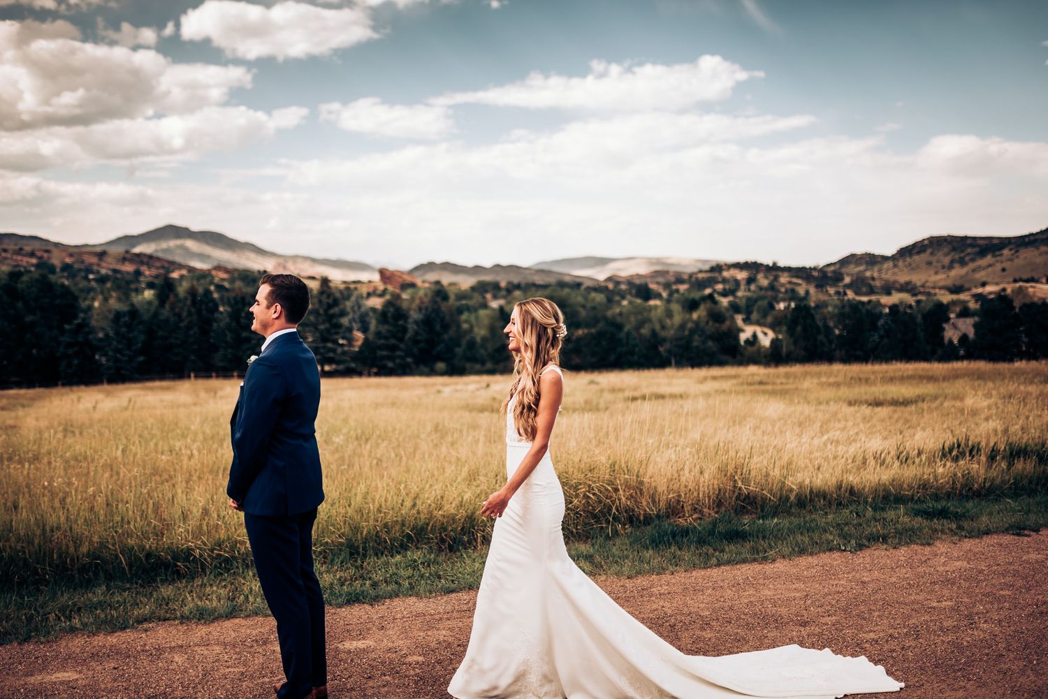 Wedding couple standing apart on dirt path with mountain landscape backdrop.