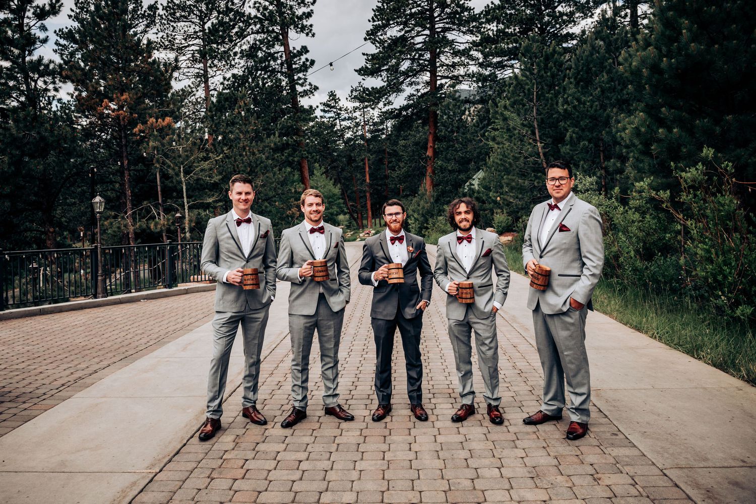 Wedding party in gray suits standing in a row on a brick pathway surrounded by pine trees.