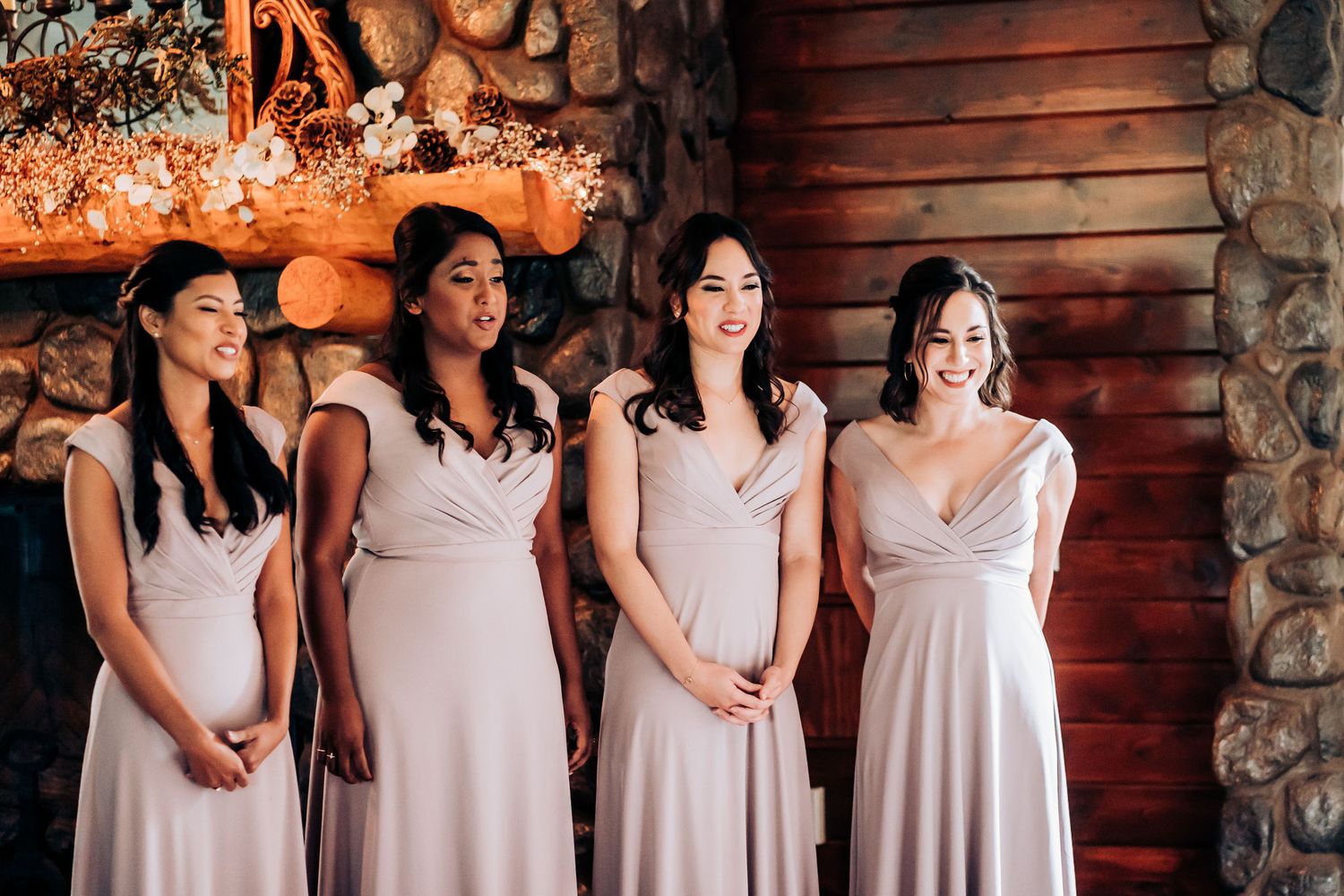 Bridesmaids in blush pink dresses stand together against rustic wooden wall.