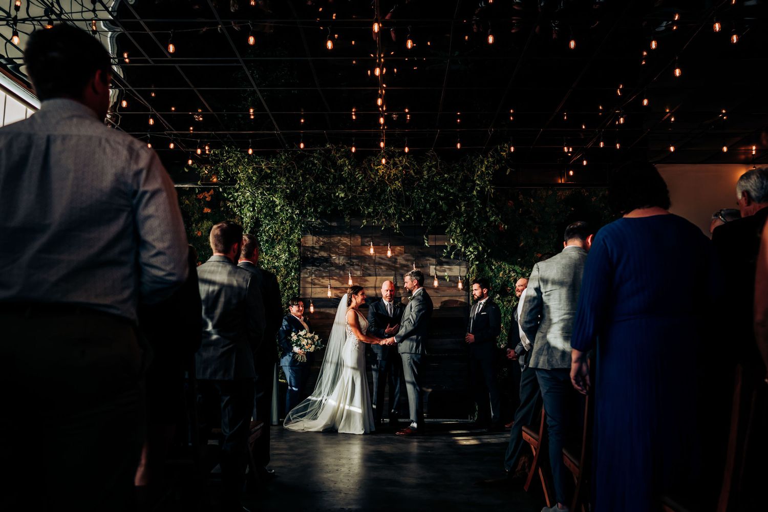Wedding ceremony in a dark venue with twinkling lights and greenery overhead as guests gather around the couple.