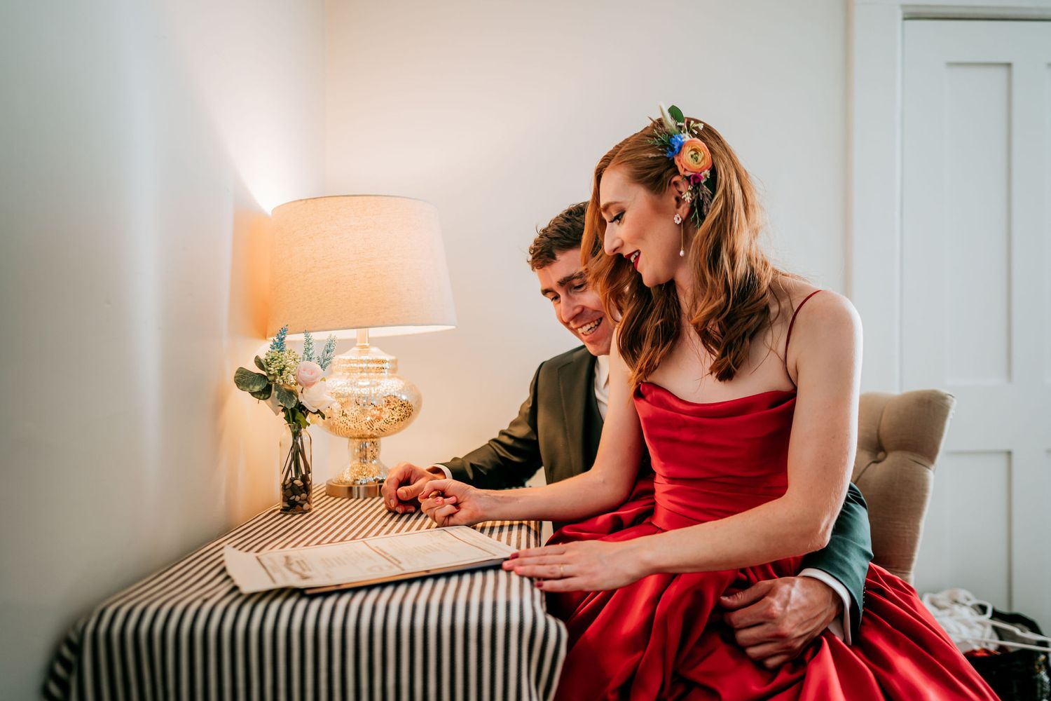 A couple shares an intimate moment while sitting together on a bed with a warm table lamp nearby.