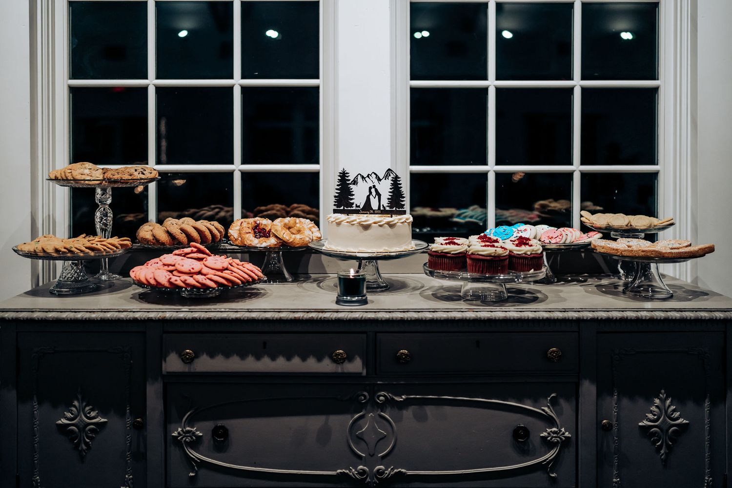Dessert table with various cakes and treats displayed against a dark window backdrop.