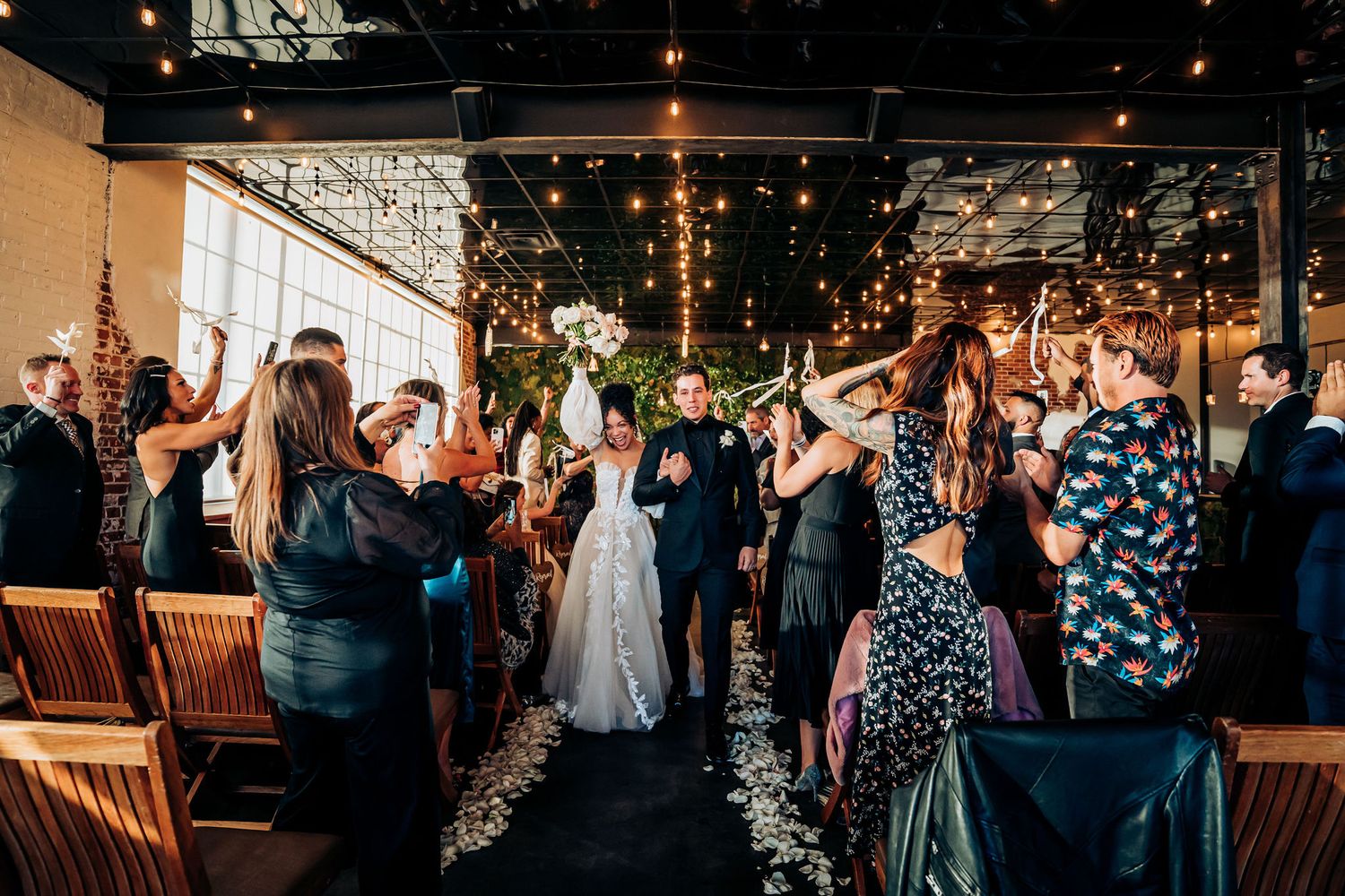 Wedding celebration with guests cheering and tossing confetti in an indoor venue with string lights.