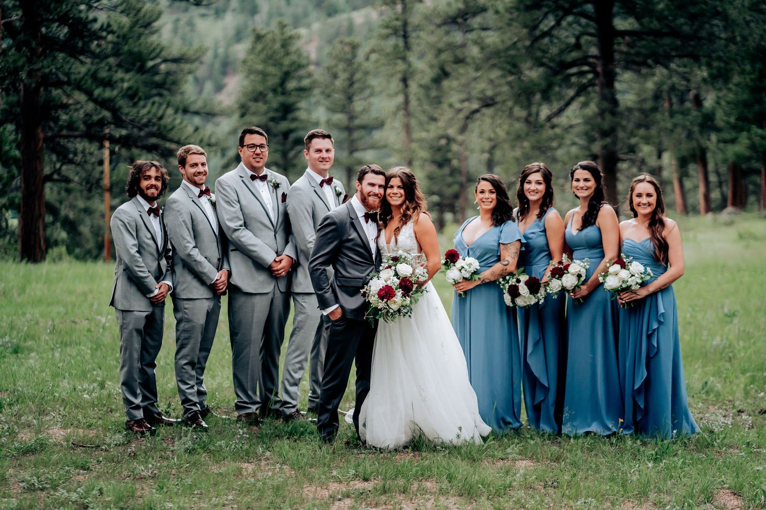 Wedding party in gray suits and dusty blue dresses pose together in a scenic mountain forest setting.