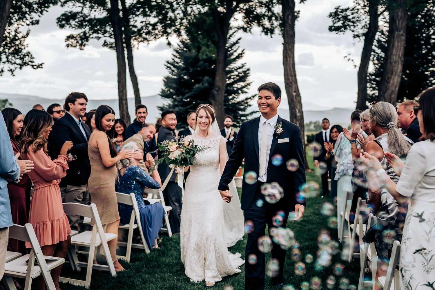 Wedding guests throw confetti as the newlyweds walk down the aisle at an outdoor ceremony.
