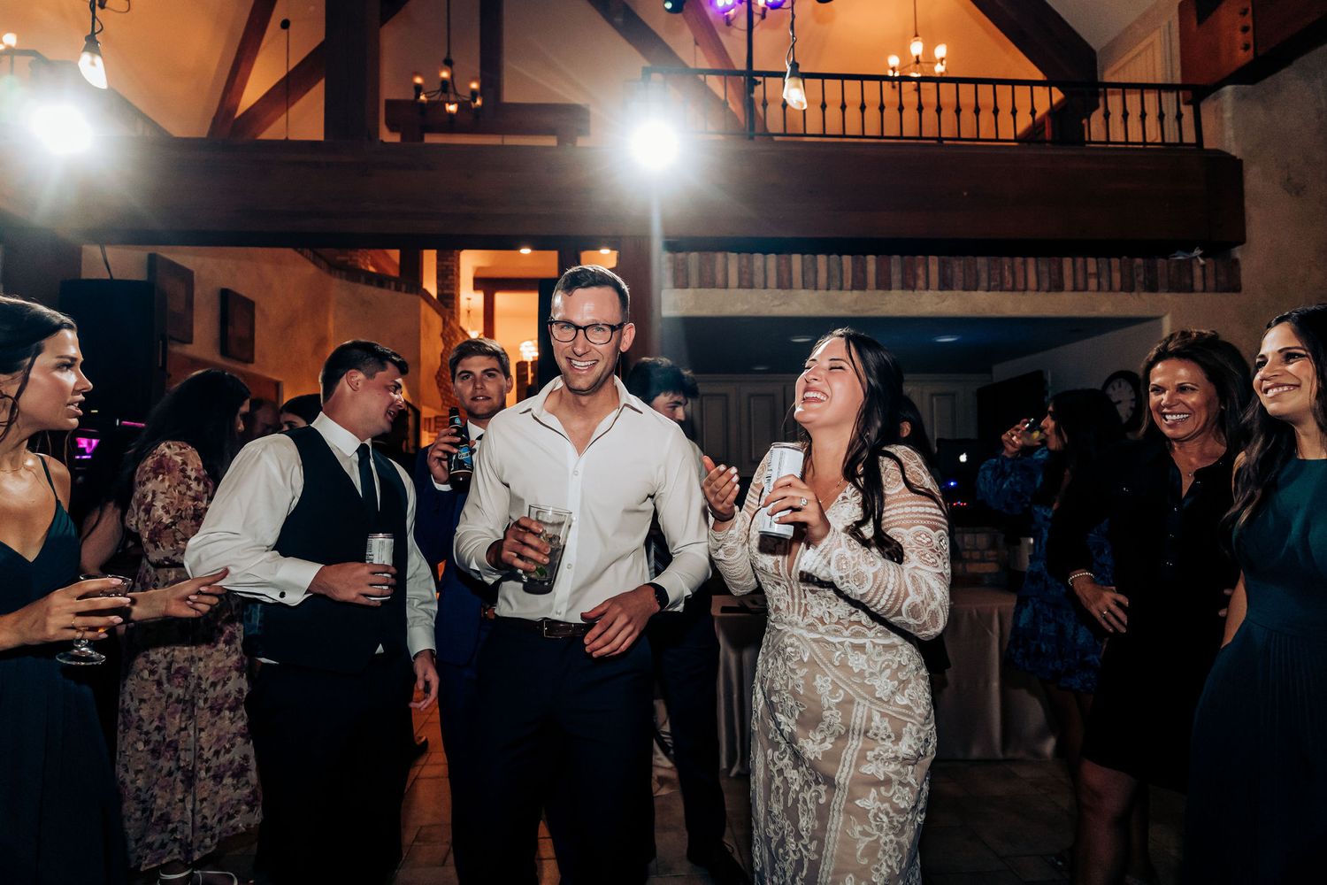Wedding reception guests mingling and drinking in rustic indoor venue with exposed beams.