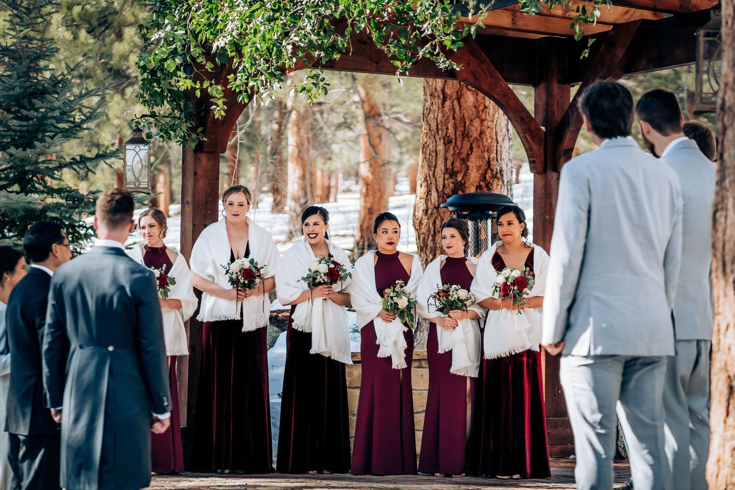 Wedding party stands under wooden arch in outdoor winter ceremony with burgundy bridesmaid dresses.