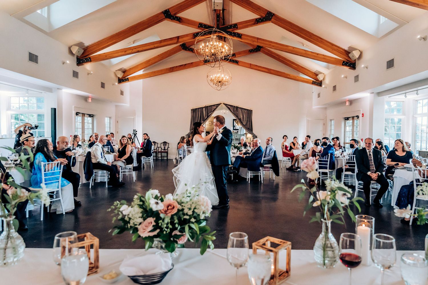 Wedding reception in bright venue with exposed wooden beams and crystal chandeliers.
