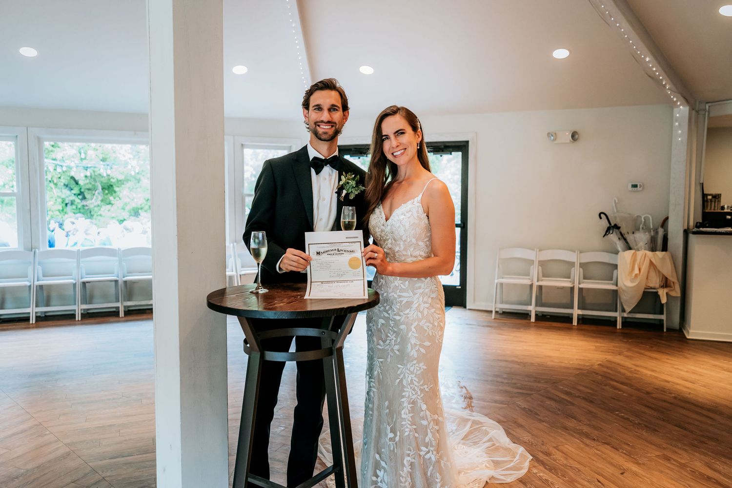 A wedding couple poses with a stack of modern white wedding cakes in an elegant reception venue.