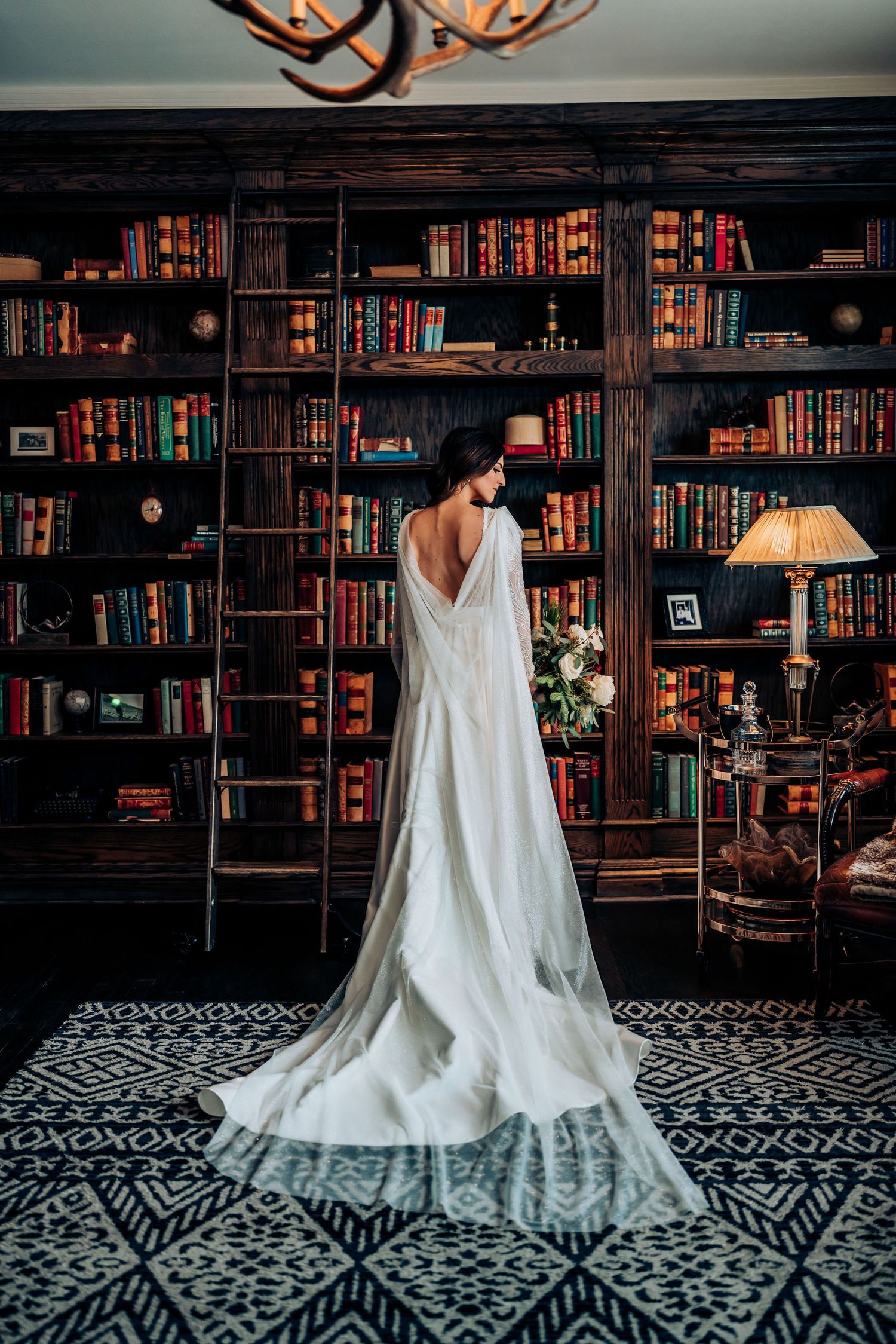 A bride in a white gown stands before a library wall with dark wooden bookshelves and patterned floor.