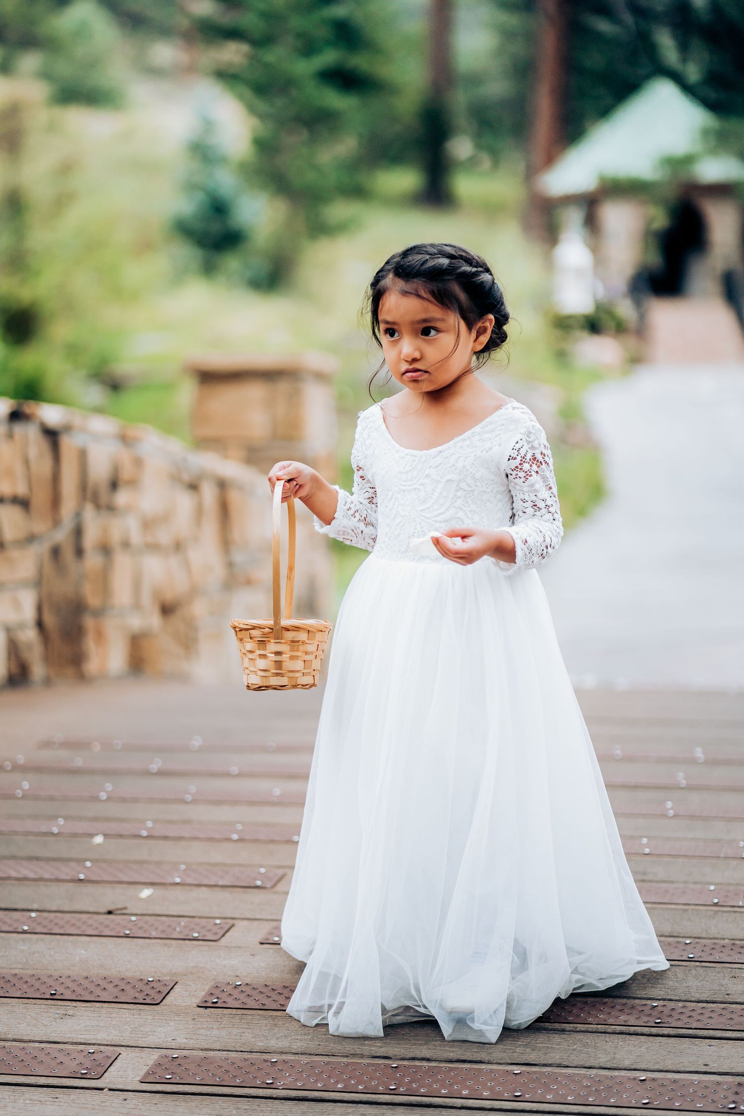 A young attendant in a white lace dress holds a basket on a wooden walkway.