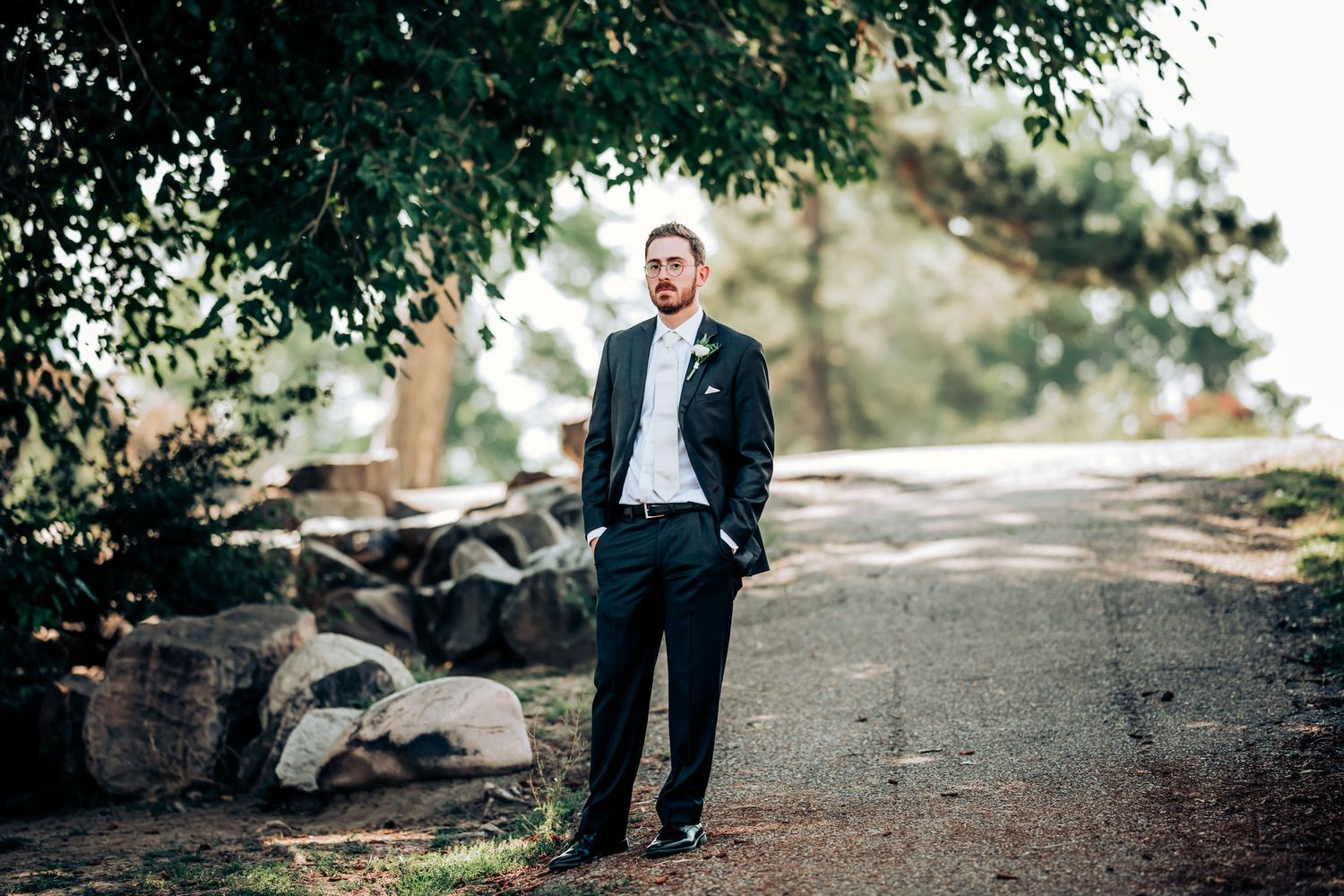 Groom in classic navy suit poses on rocky garden path under tree canopy.