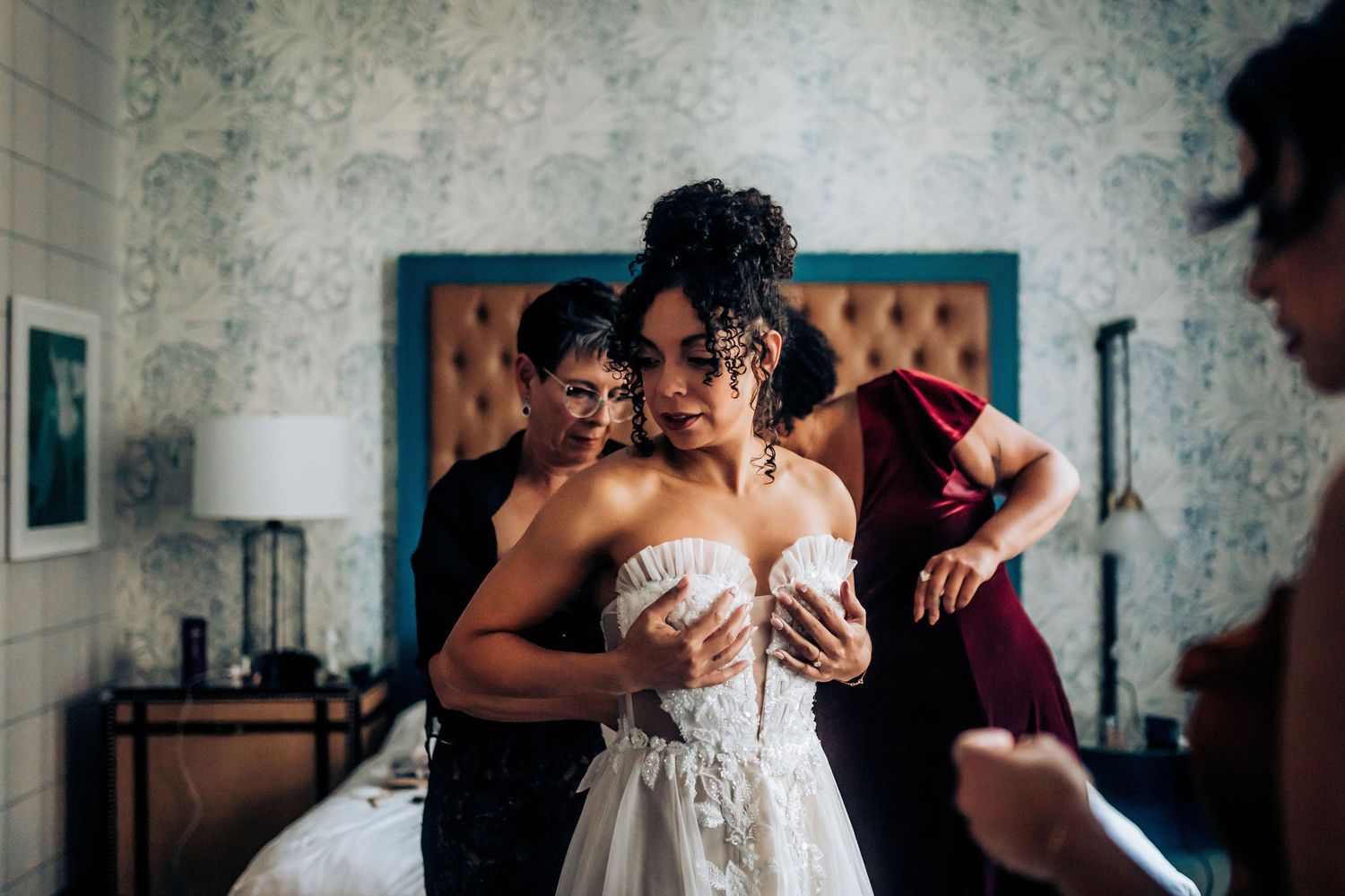 A bride in a white strapless gown gets help getting dressed in a room with floral wallpaper and orange headboard.