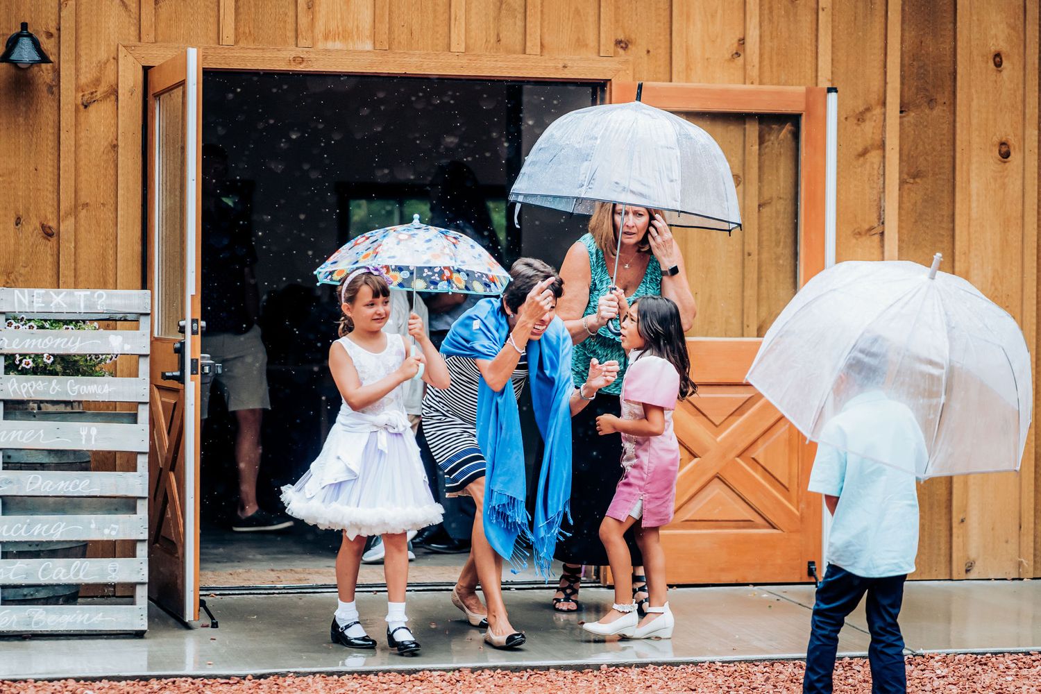 A group shelters under white umbrellas outside a rustic wooden barn entrance.