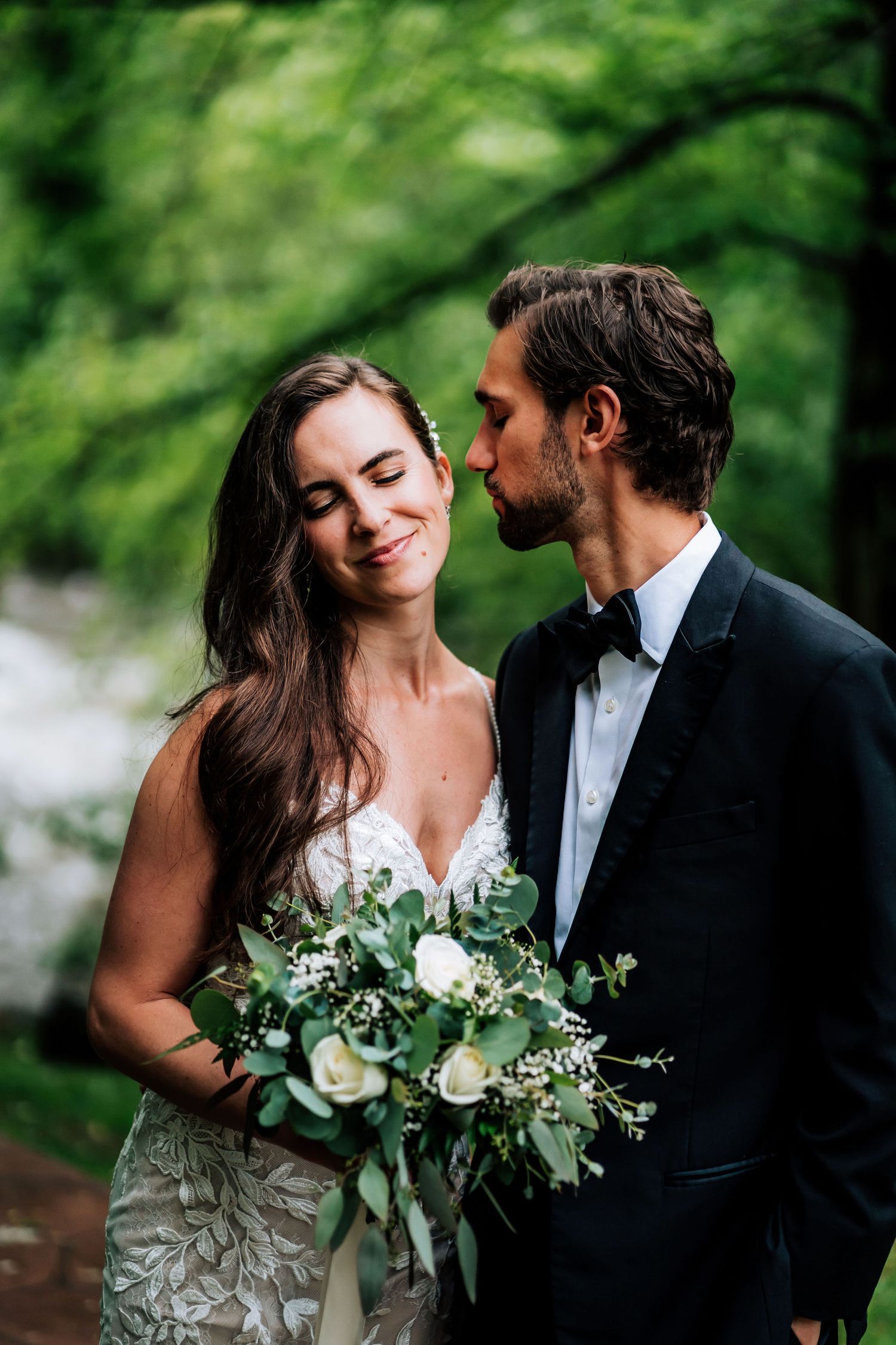 A romantic moment between newlyweds with a lush green forest backdrop and white floral bouquet.