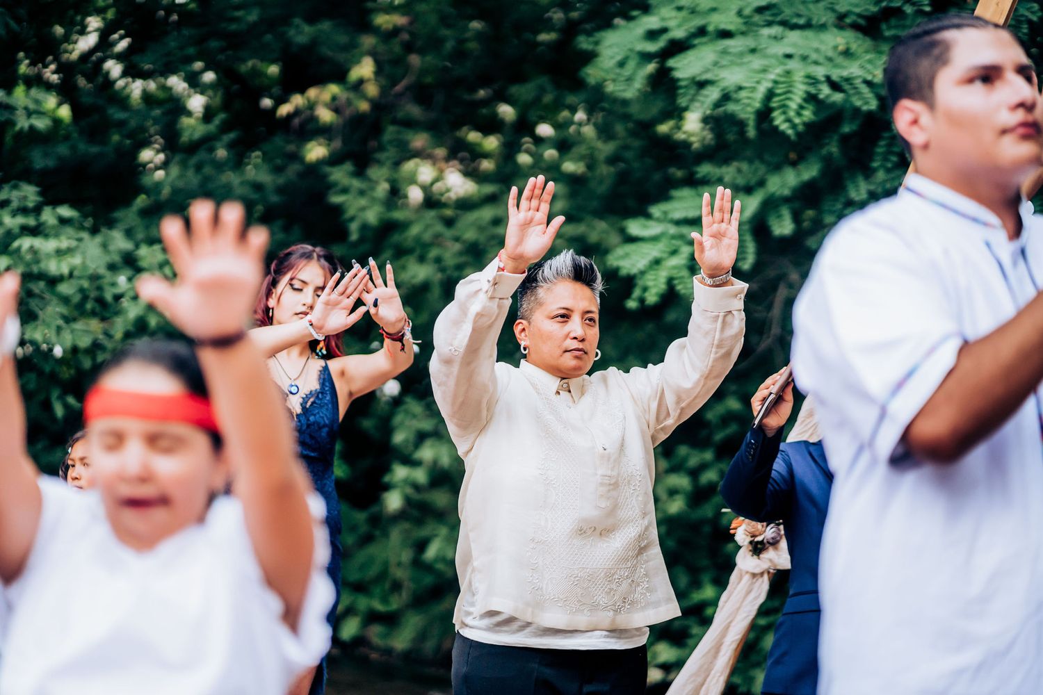 Group of wedding attendees raising their hands during outdoor celebration with trees in background.