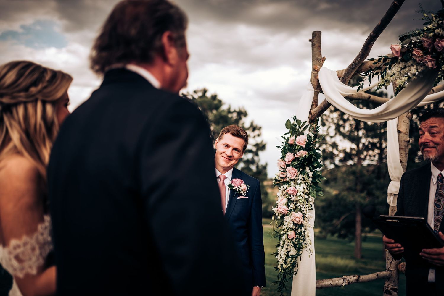 Wedding ceremony under a rustic arch decorated with flowers and draped fabric in front of trees.