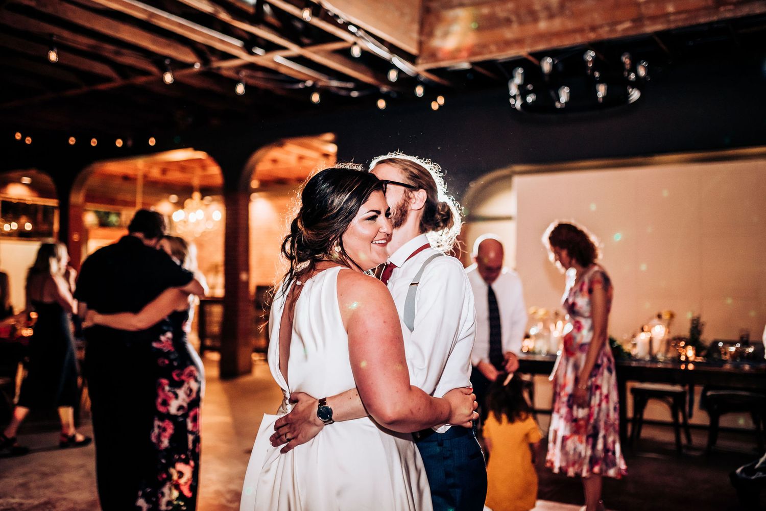 Couple embraces during a tender moment on their wedding day in a rustic venue with string lights.