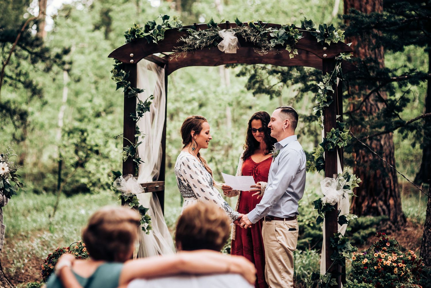 A wedding ceremony under a flower-decorated wooden arch in a pine forest.