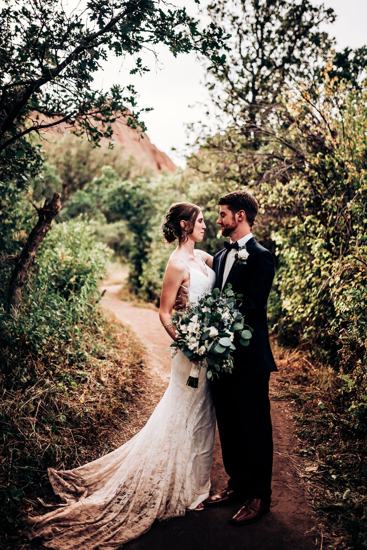 Couple in wedding attire shares intimate moment on forest path with flowing lace dress.