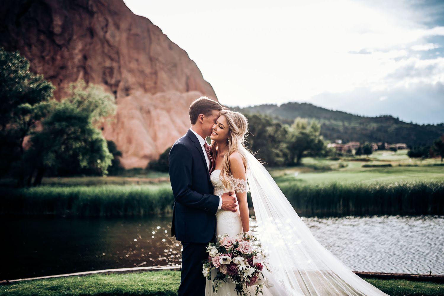 Wedding couple shares intimate moment against dramatic red rock formation and lake backdrop at sunset.