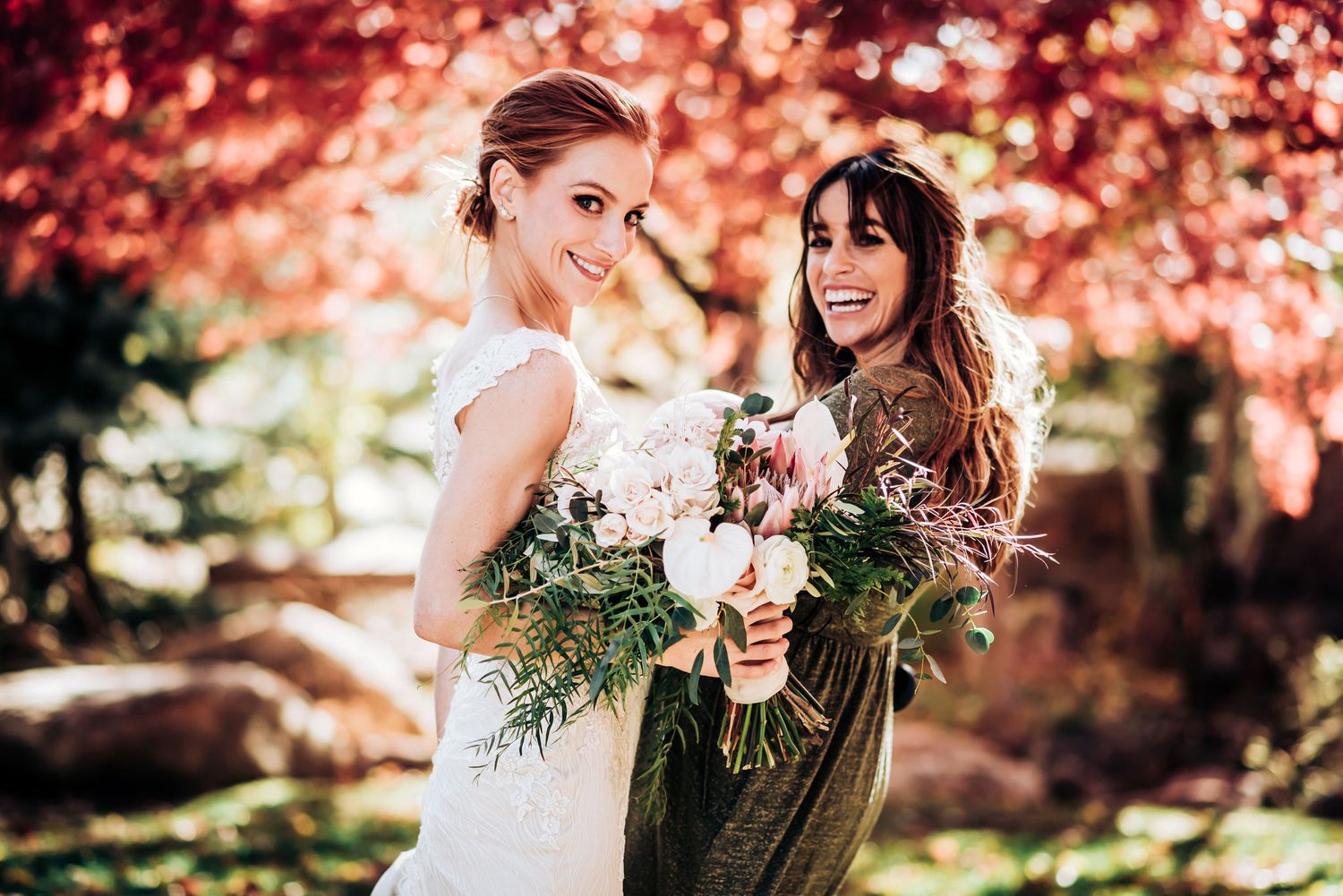 Two friends pose with wedding bouquet against stunning fall foliage backdrop.