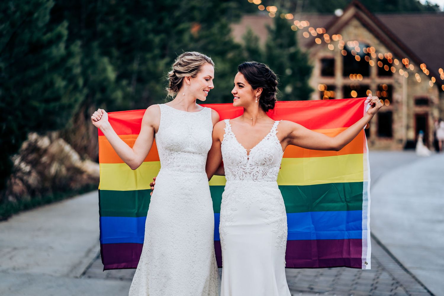 Two people in white wedding dresses hold a rainbow pride flag at an outdoor wedding venue celebration.