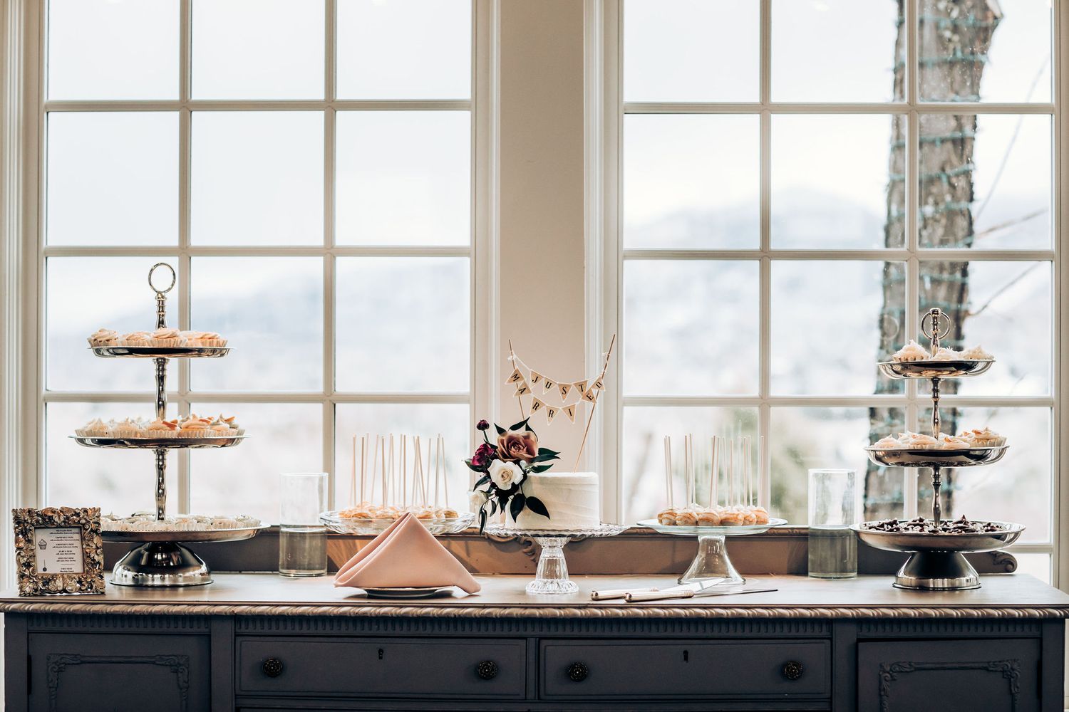 Elegant dessert and appetizer buffet display on black sideboard with tiered stands near bright windows.