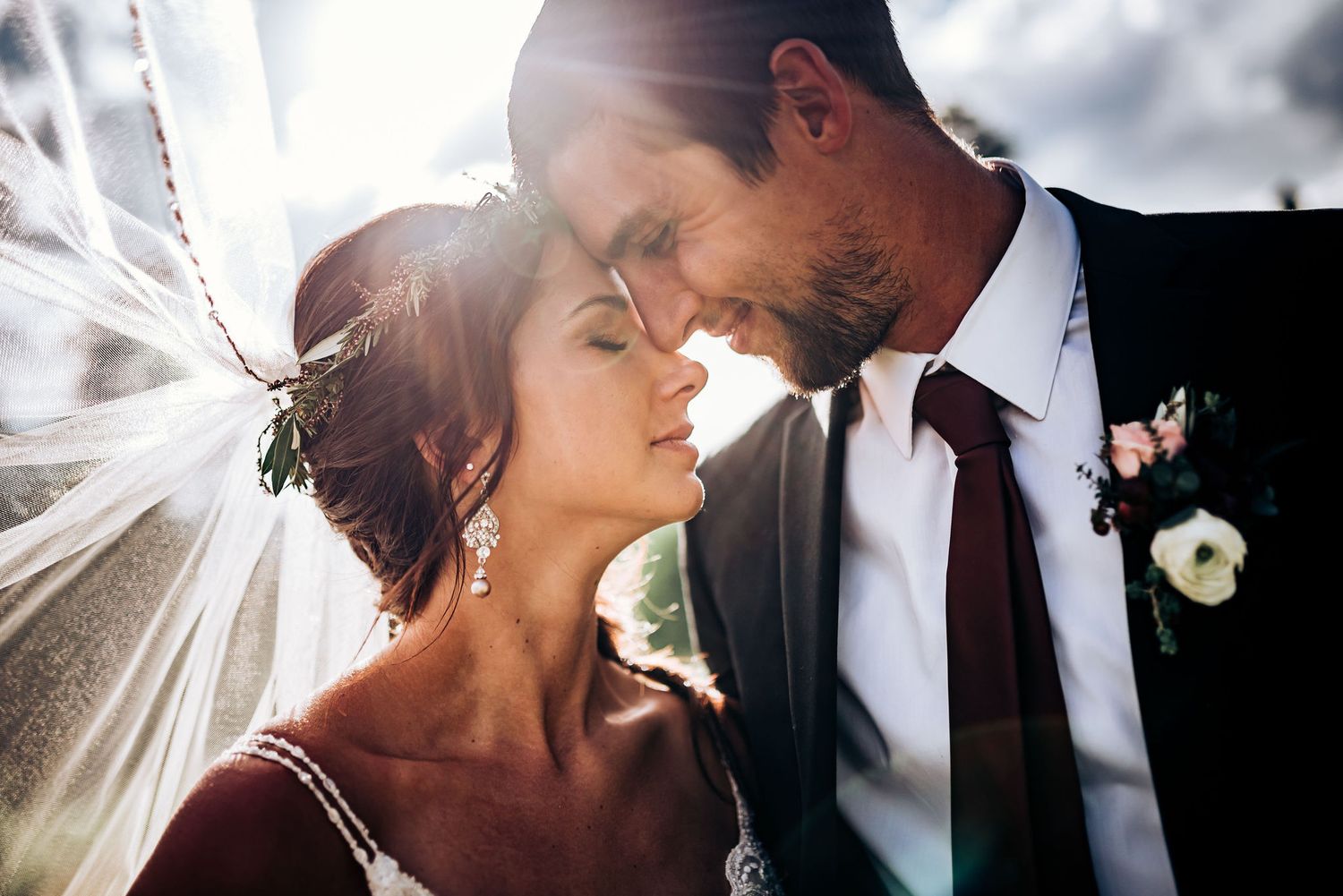 Sunlit wedding portrait shows couple in tender embrace with flowing veil and classic formal wear.