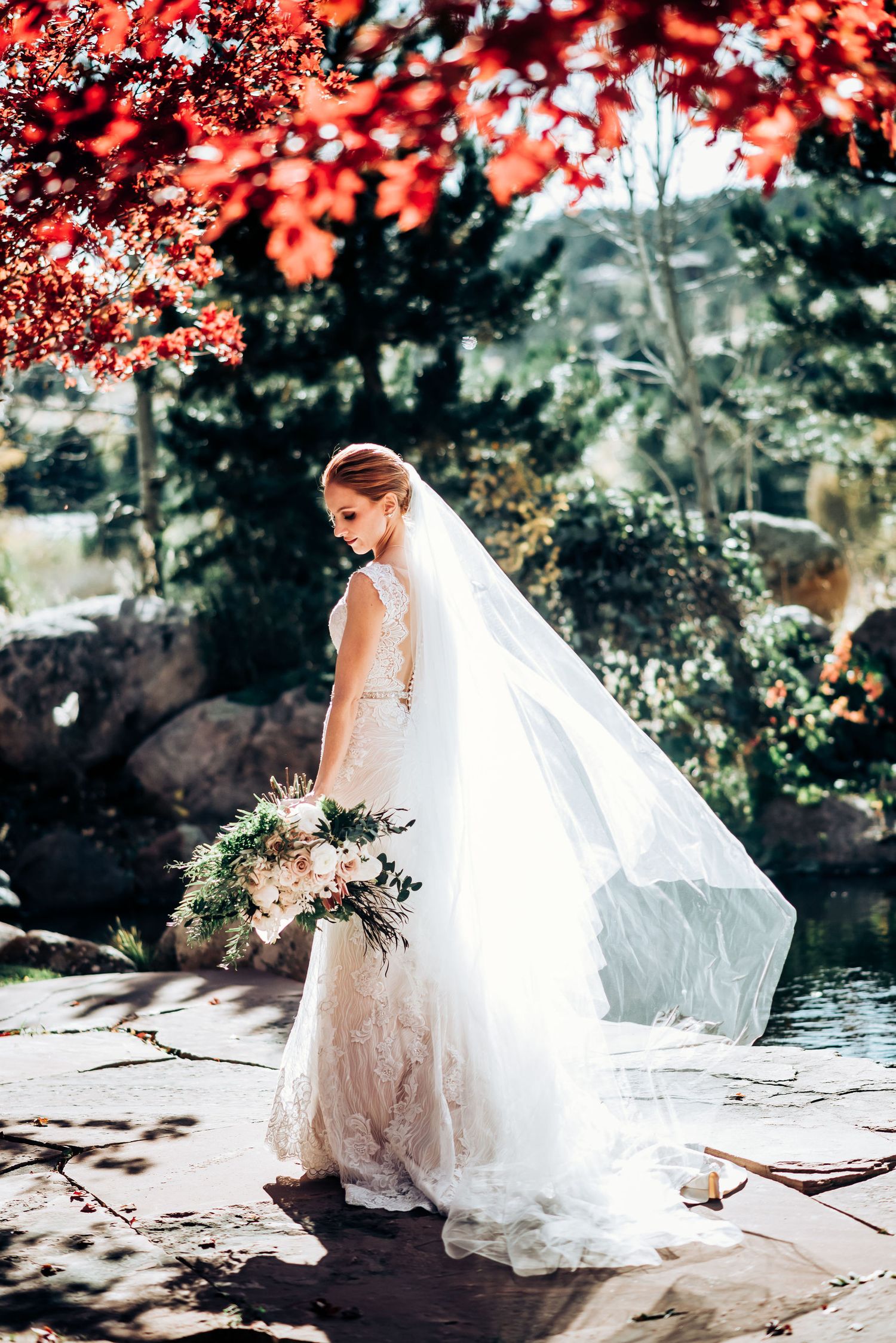 Bride in lace gown with flowing veil poses against backdrop of red autumn leaves.