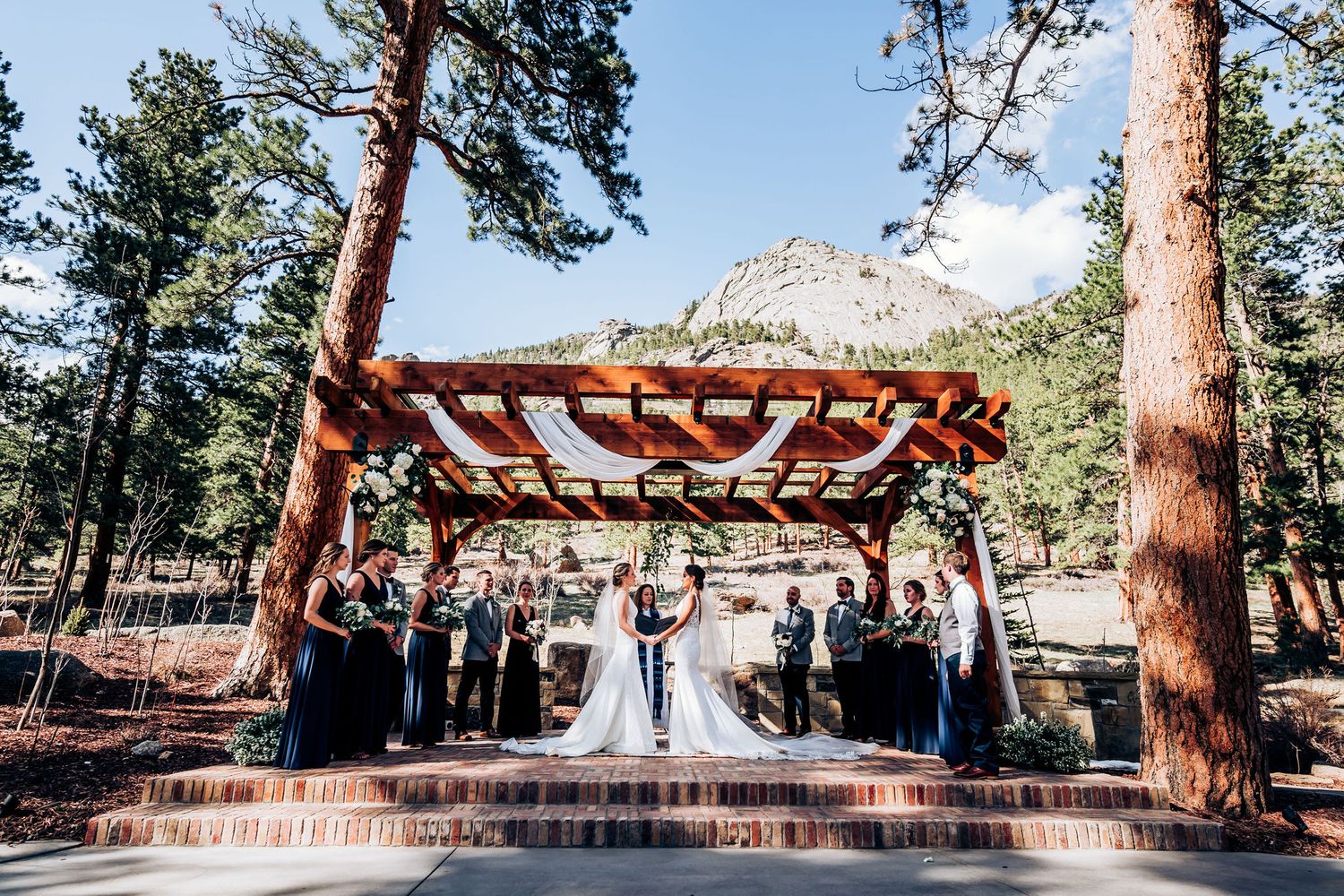 An outdoor wedding ceremony beneath a rustic wooden pergola with mountain views and tall pine trees.