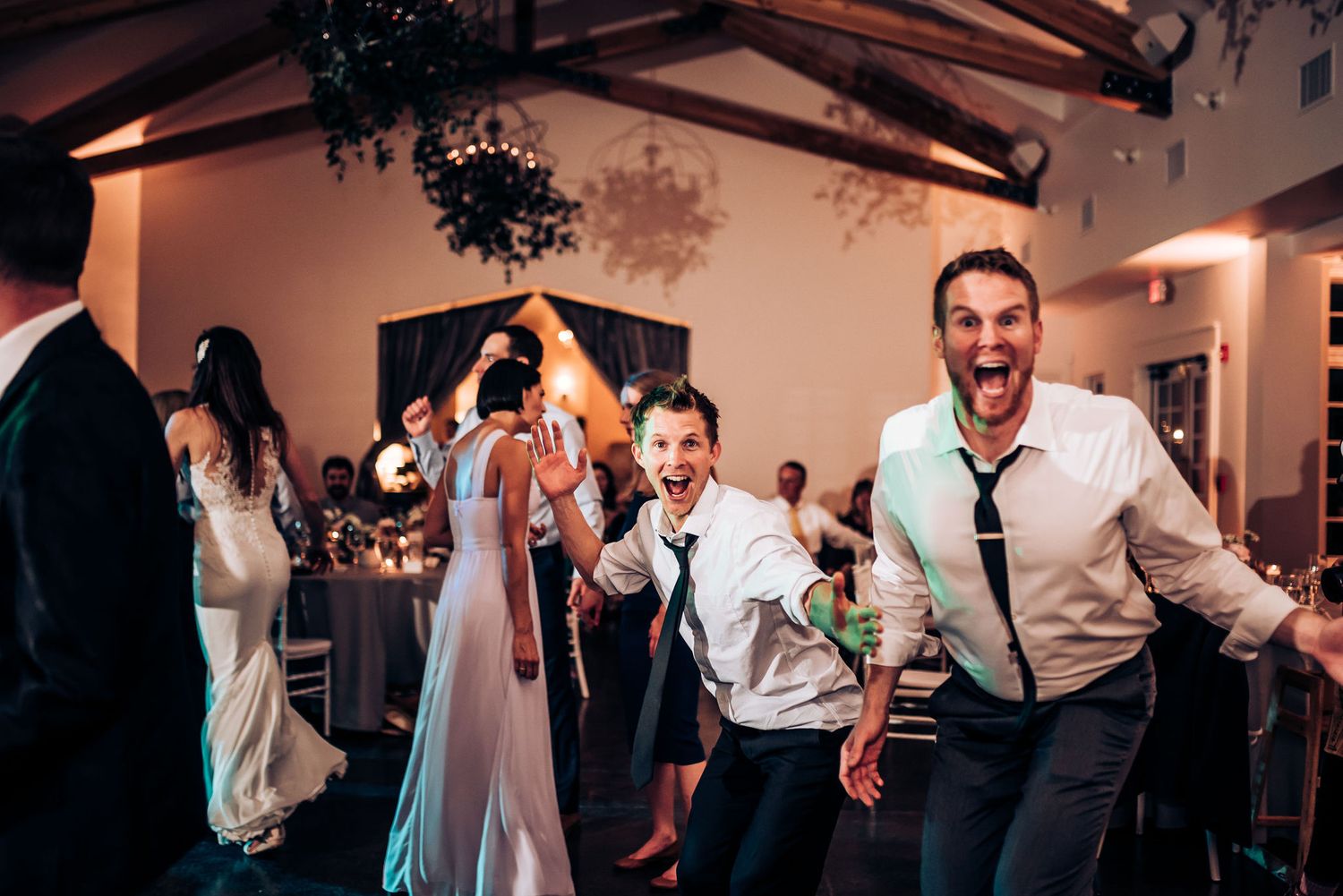 Energetic wedding reception dance floor with guests celebrating under wooden beams and decorative lighting.