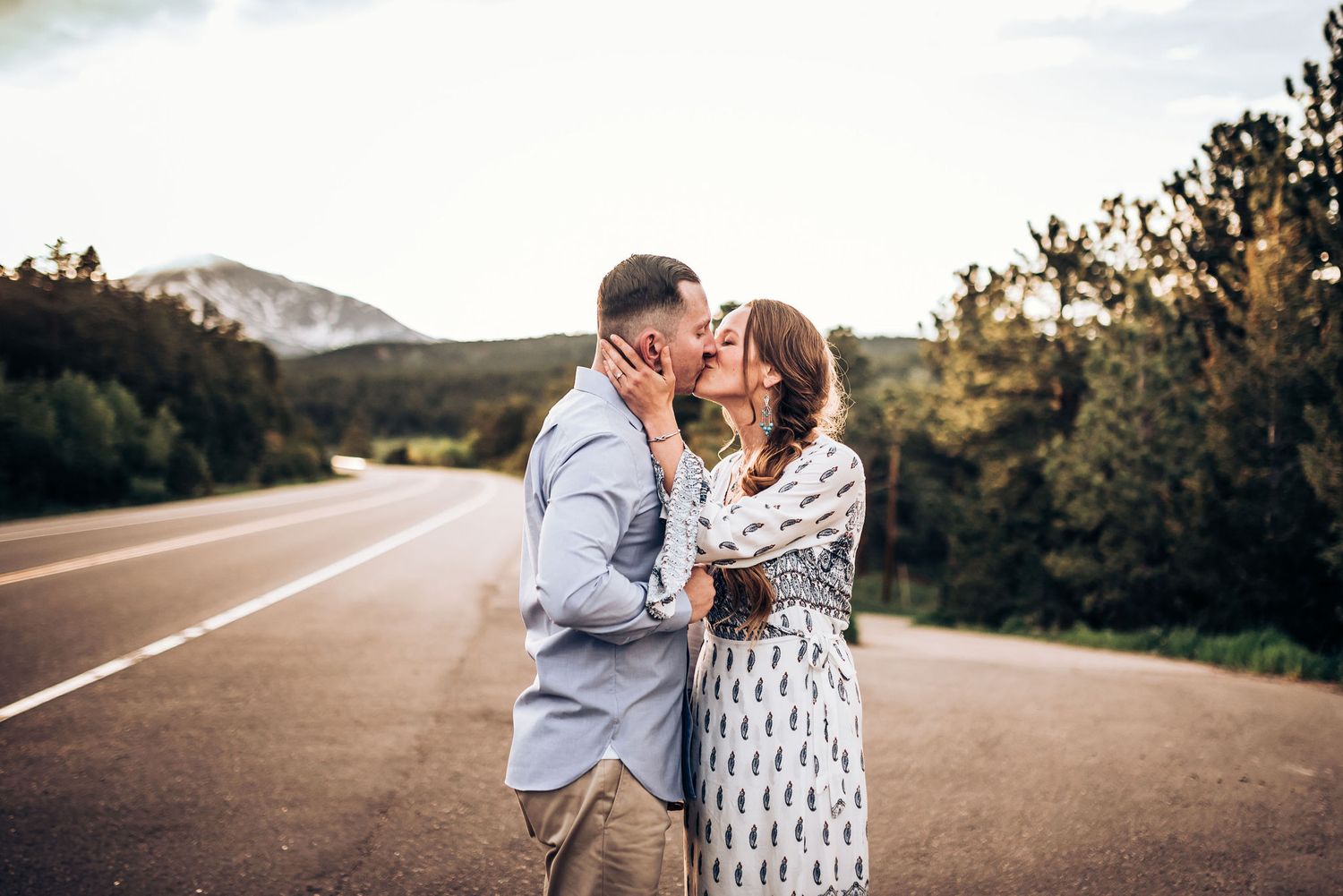 A romantic sunset photoshoot on a mountain road features a couple embracing in coordinated grey and white outfits.