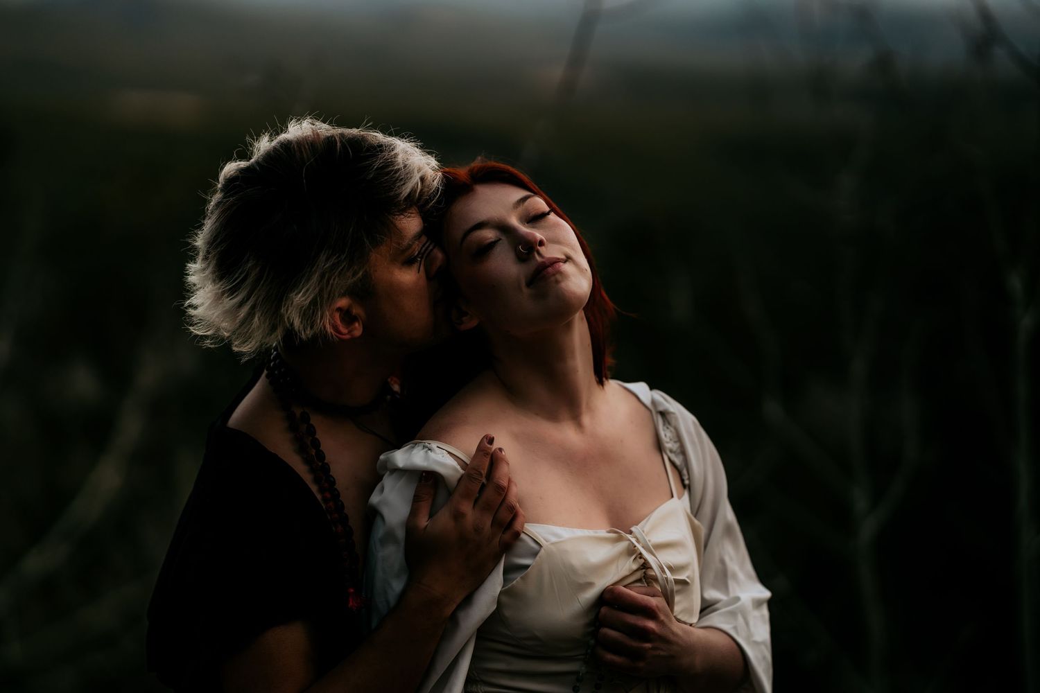 A couple shares an intimate embrace in a dark, moody outdoor setting with dramatic lighting and shadows.