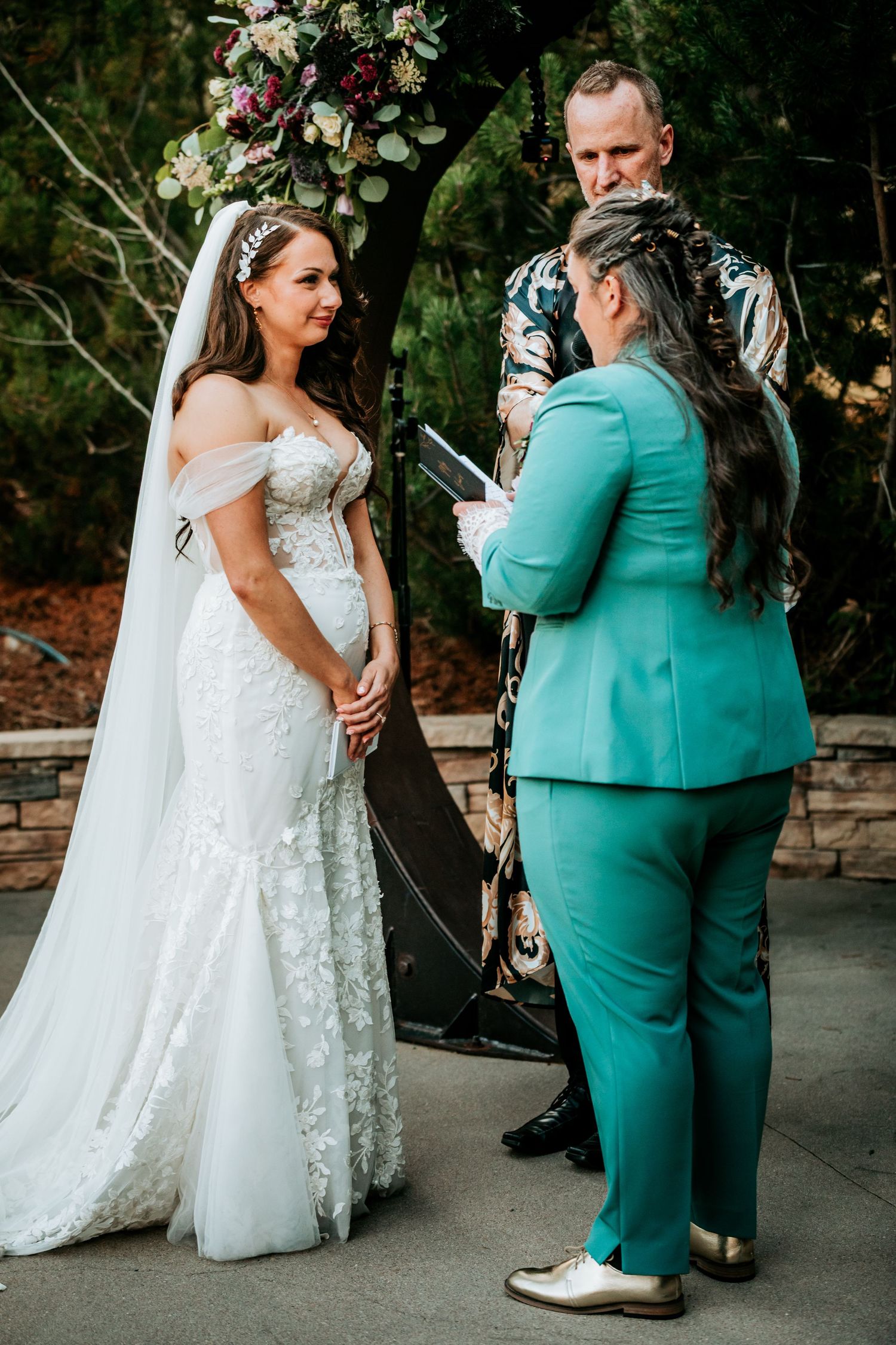 Bride in white lace gown and partner in teal suit exchanging vows at outdoor ceremony with officiant.