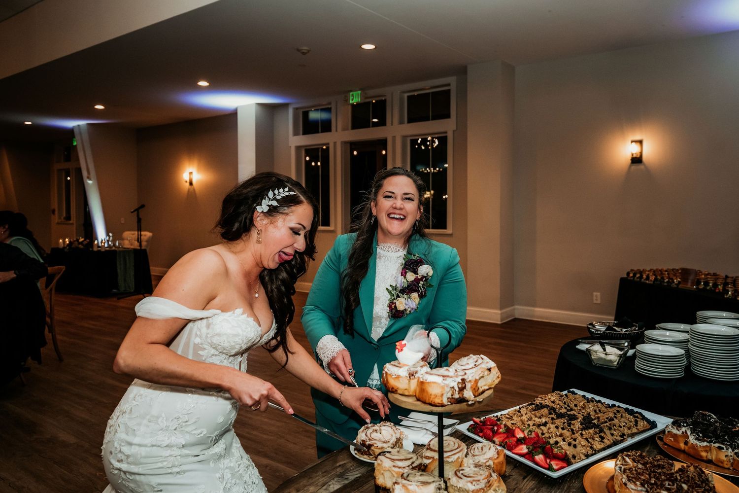 Two brides laugh while cutting their wedding cake at a reception dessert table filled with pastries and fruit.