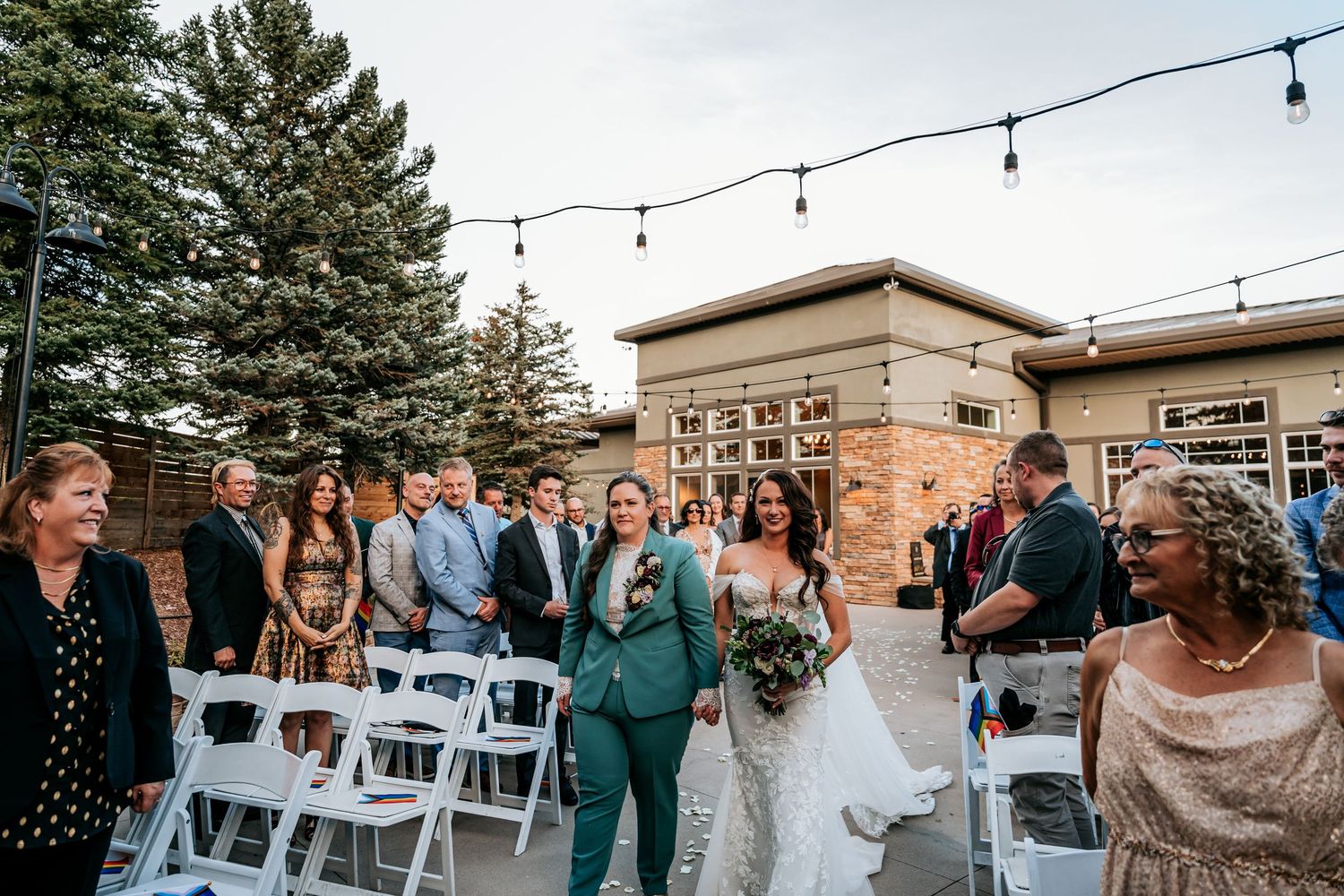 Two brides walk joyfully down aisle after LGBTQ wedding ceremony outdoors.
