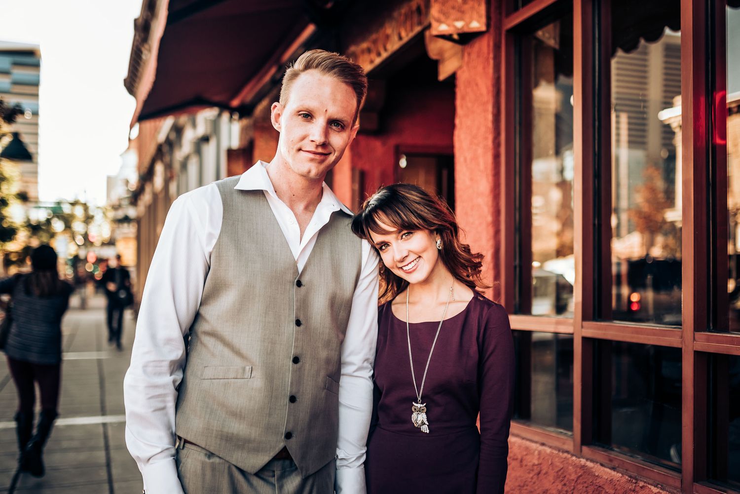 A stylish couple poses together outside urban storefronts wearing coordinated formal attire.