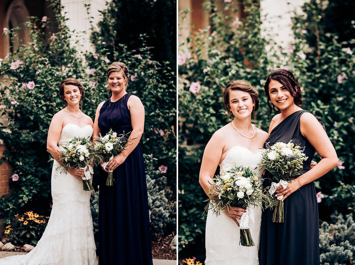 A wedding photo showing two people in navy and white dresses posing in front of blooming rose bushes.
