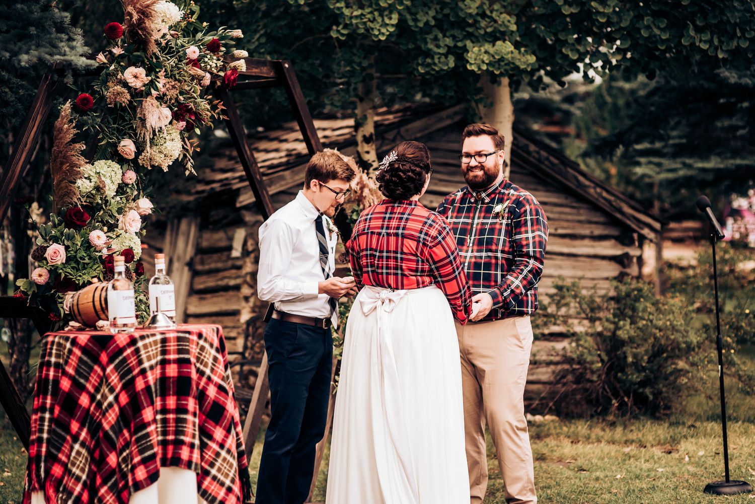 Wedding ceremony participants gather near a rustic cabin decorated with floral arrangements.