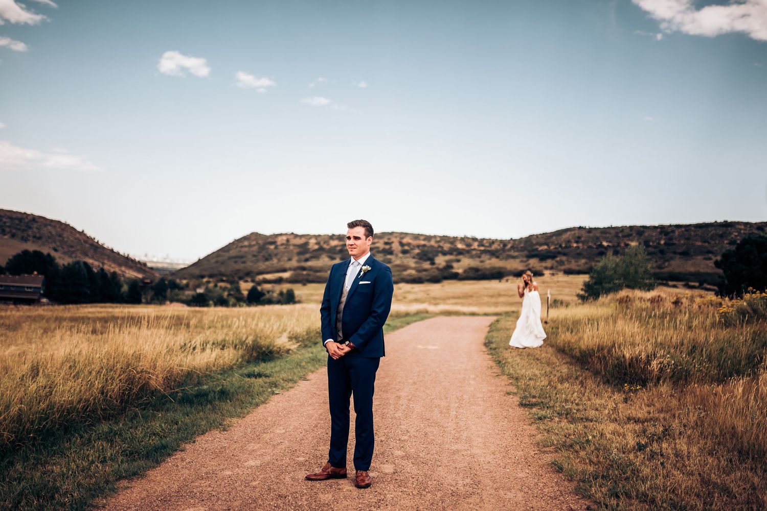 Wedding couple separated on dirt path with mountains and golden fields in background.