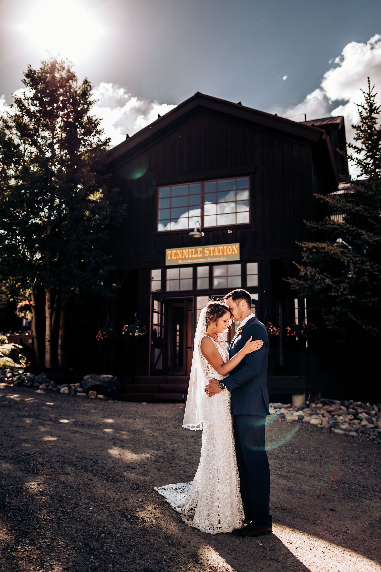 A romantic wedding couple embraces outside a rustic wooden building with sun flares in the background.