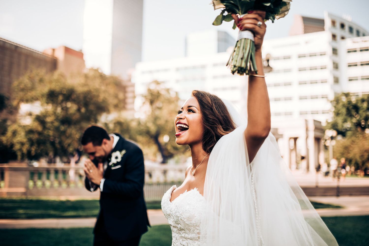 A bride celebrates joyfully outside with raised bouquet while silhouetted figure stands in background.